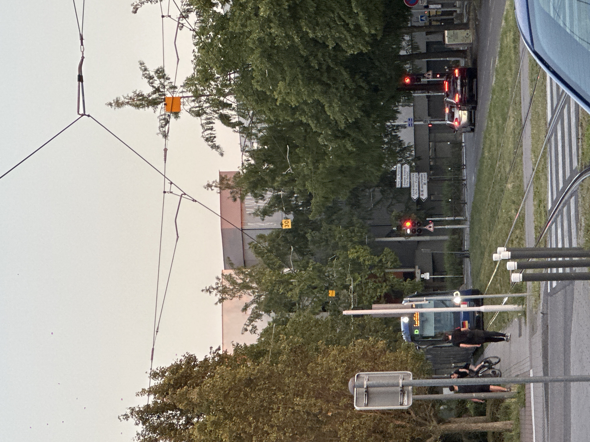 This image depicts a street scene in Strasbourg, France, with a tram visible on the tracks. In the foreground, there are tram tracks and a pedestrian crossing. To the left, a blue tram is stopped, with several people nearby. One person is walking alongside the tram, another is on a bicycle, and a third is standing. Further down the street, there are trees, buildings, and traffic lights. Two cars are stopped at a red traffic light. There are overhead wires for the tram. The sky is overcast, suggesting it is either early morning or late evening. Signs indicating directions are visible on a pole. One sign reads "Parc de Poteries", suggesting this area may be near a park with that name in Strasbourg.