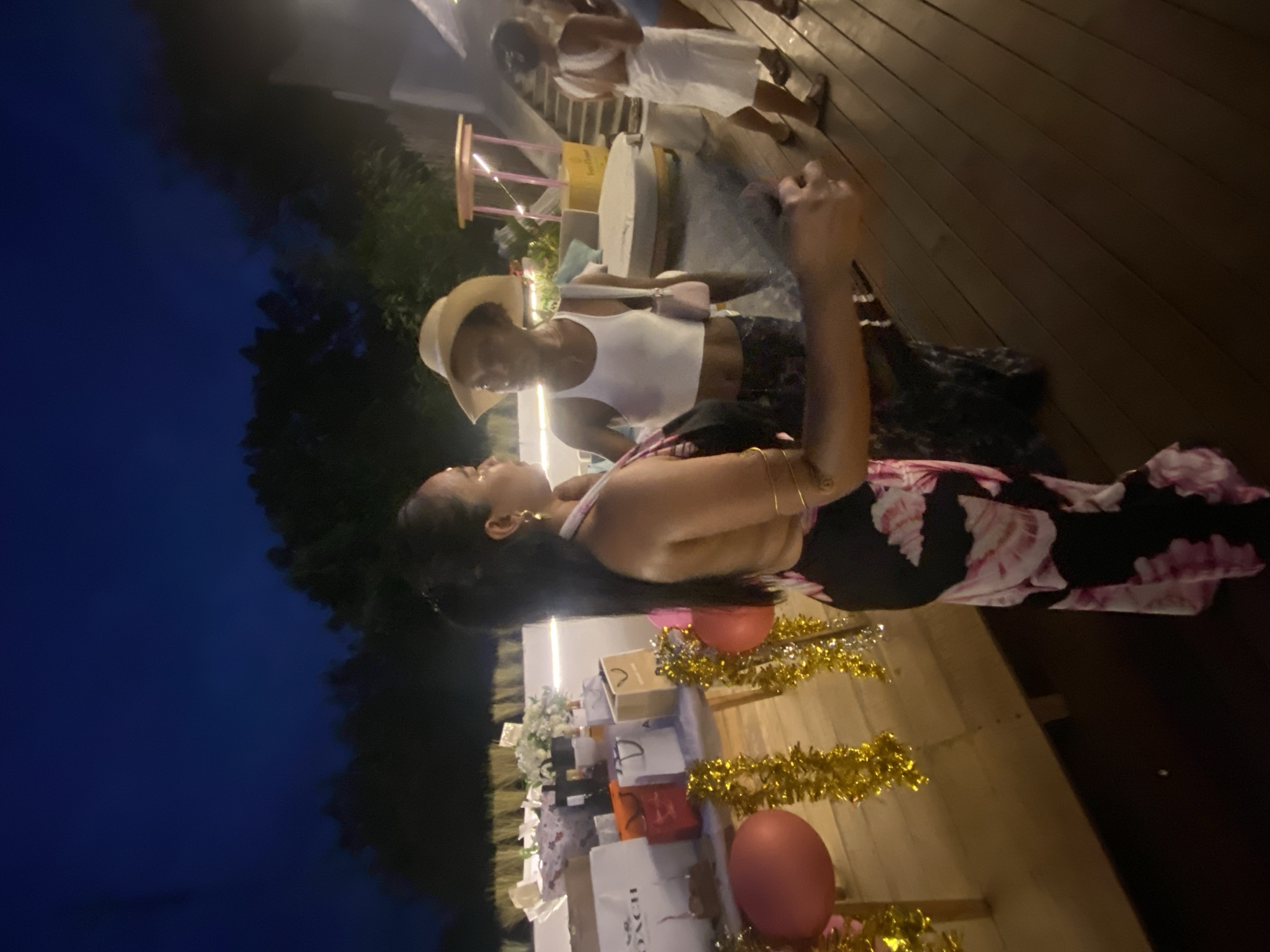 Two women are standing on a wooden deck at night, possibly at an outdoor event or party. The woman in the foreground is wearing a black dress with a floral print and has her hair down. She is looking to her left, towards the other woman. She is wearing gold hoop earrings and gold bracelets. The second woman, to her left, is wearing a white tank top and a straw hat. She is also looking at the first woman. There are several gift bags and balloons on a table in front of them, suggesting a celebration. The background shows trees and a dark blue night sky, indicating it is nighttime. The location is identified as Nusa Penida, Indonesia.