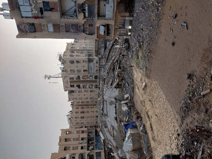 Heavily damaged buildings stand amidst rubble and debris. The structures show extensive destruction, with multiple floors exposed and facades torn away. A sandy or gravelly terrain surrounds the ruins, littered with scattered debris. The sky is overcast and pale. The scene conveys devastation and destruction.