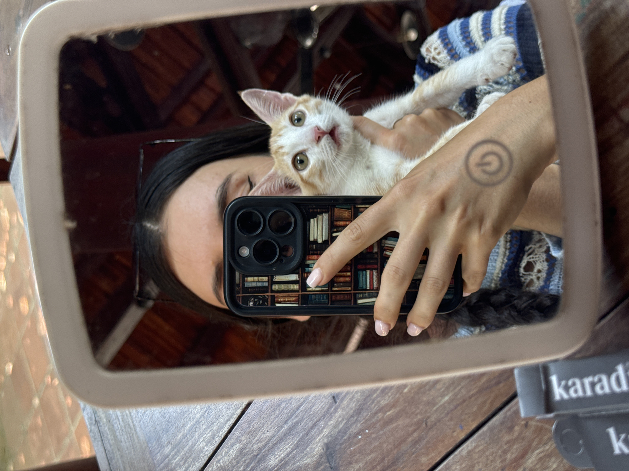 In a selfie reflected in a mirror, a person holds a white and orange kitten. The person's face is partially obscured by a smartphone, which displays a book-themed phone case. The person has long dark hair and a tattoo of a power button symbol on their left forearm. The kitten has wide, green eyes and is looking directly at the camera. The reflection also shows a striped sweater worn by the person. The background suggests an outdoor setting, possibly a cafe or restaurant, with wooden tables and roofing structures visible in the mirror's reflection, characteristic of Mũi Né, Vietnam.