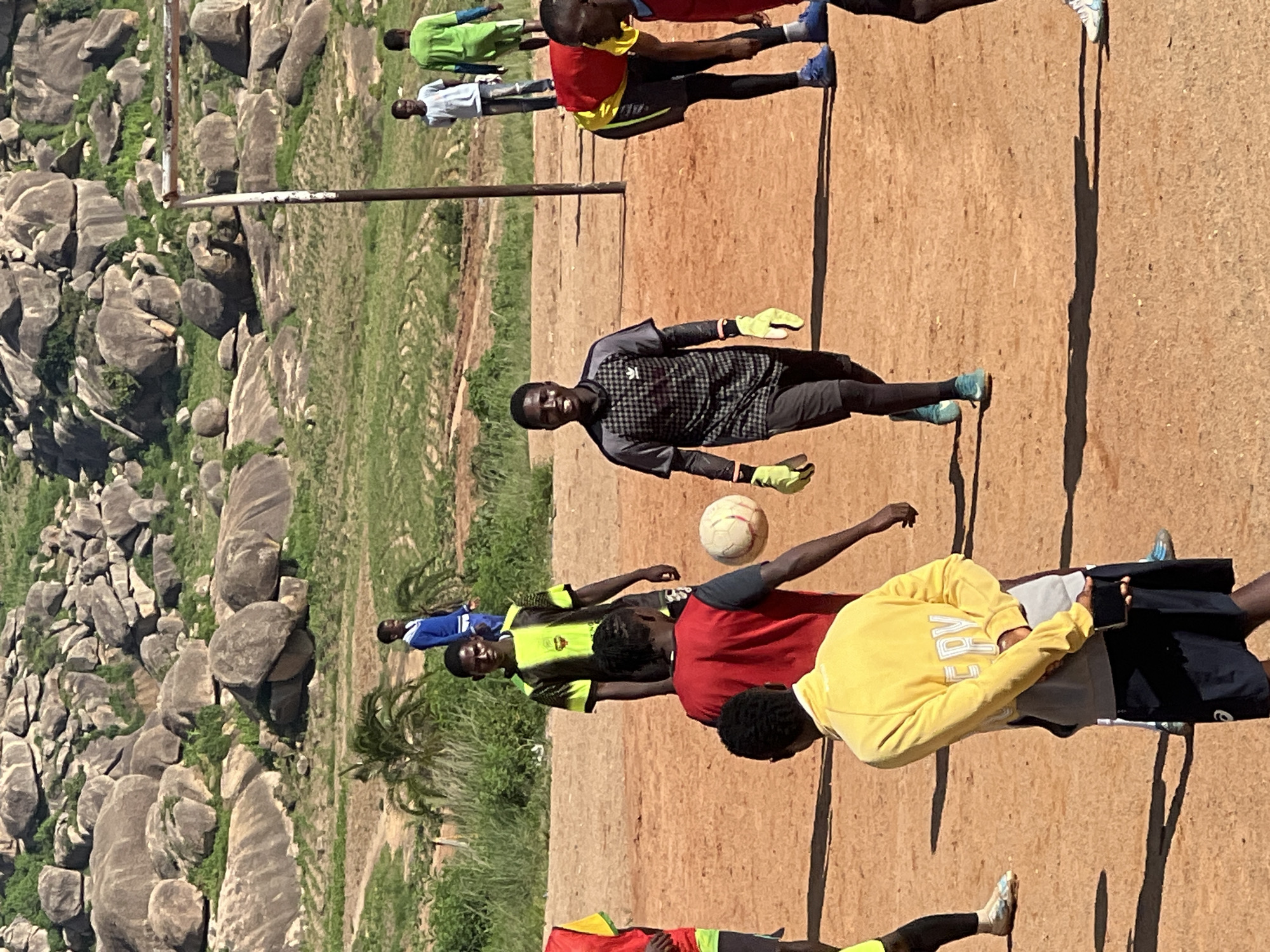 The image depicts a group of young men playing football on a dirt field with a rocky, grassy hillside in the background, possibly in Jos, Nigeria. In the foreground, a goalkeeper in a grey and black checkered jersey and yellow gloves is smiling as he walks towards the viewer. To his left, another player in a red vest is partially visible. Behind them, a player in a yellow jacket is holding a phone and facing away. Further back, several other individuals are scattered around the field, some in athletic wear, suggesting a casual game. A goalpost is visible on the right side of the frame. The environment appears to be arid, with sparse vegetation and large rock formations characteristic of some areas in Nigeria.