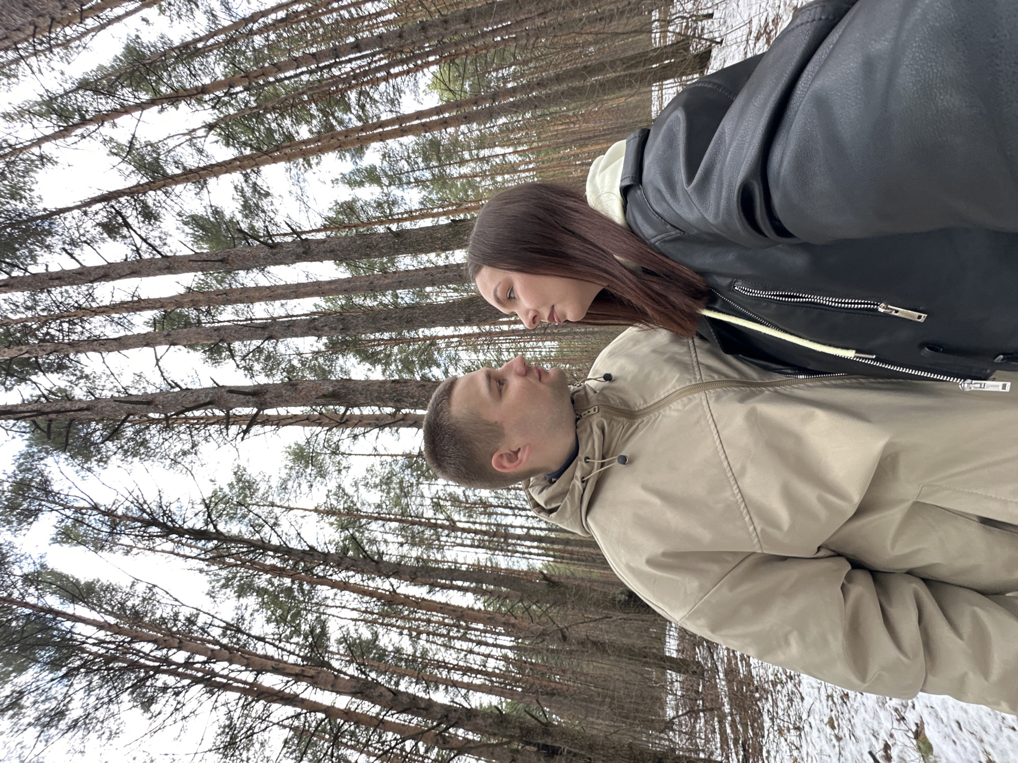 The image shows a couple standing in a forest. The man is wearing a beige hooded jacket and the woman is wearing a black leather jacket. They are looking at each other. The background consists of tall trees. The ground appears to be covered with some snow. Based on geo-location data, the location is likely in the city of Chaikovsky, Russia.