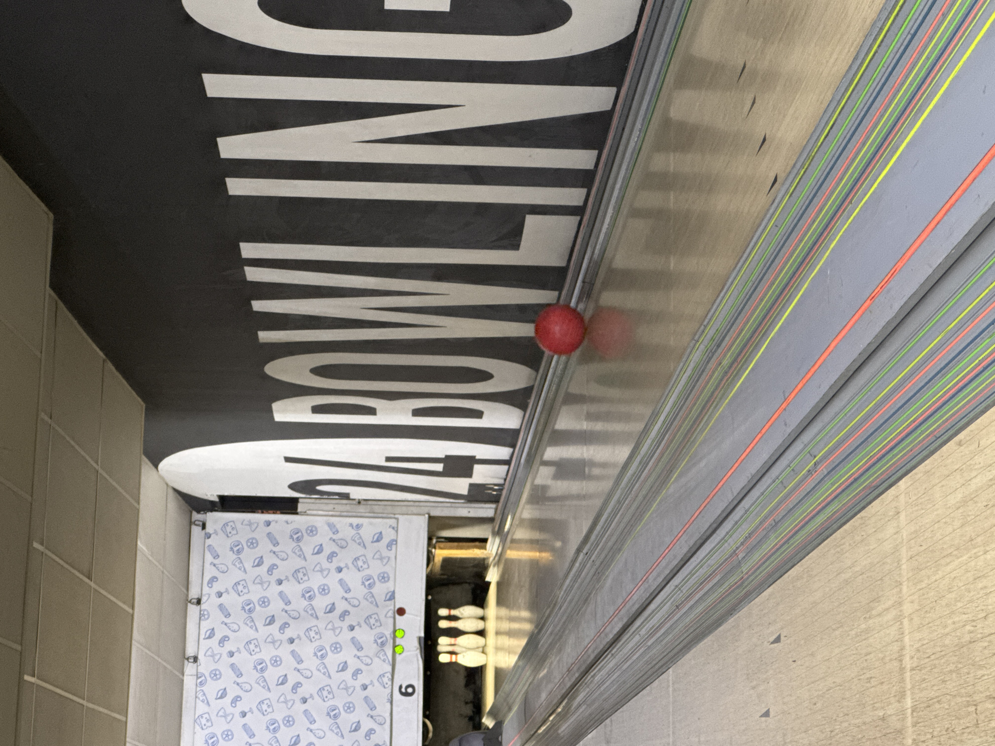 The image depicts a bowling alley. The ceiling is black with the words "24 BOWLING" in large white letters. A red bowling ball sits on the ball return, a gray metal structure with colored stripes. A lane with bowling pins is visible in the background. The number "6" is visible on a side panel near a dispenser-like area with a white and blue patterned design, possibly for snacks or balls. The floor appears to be tiled in a light color. There is no mention or indication of the location being in Kharkiv, Ukraine. No people are clearly visible.