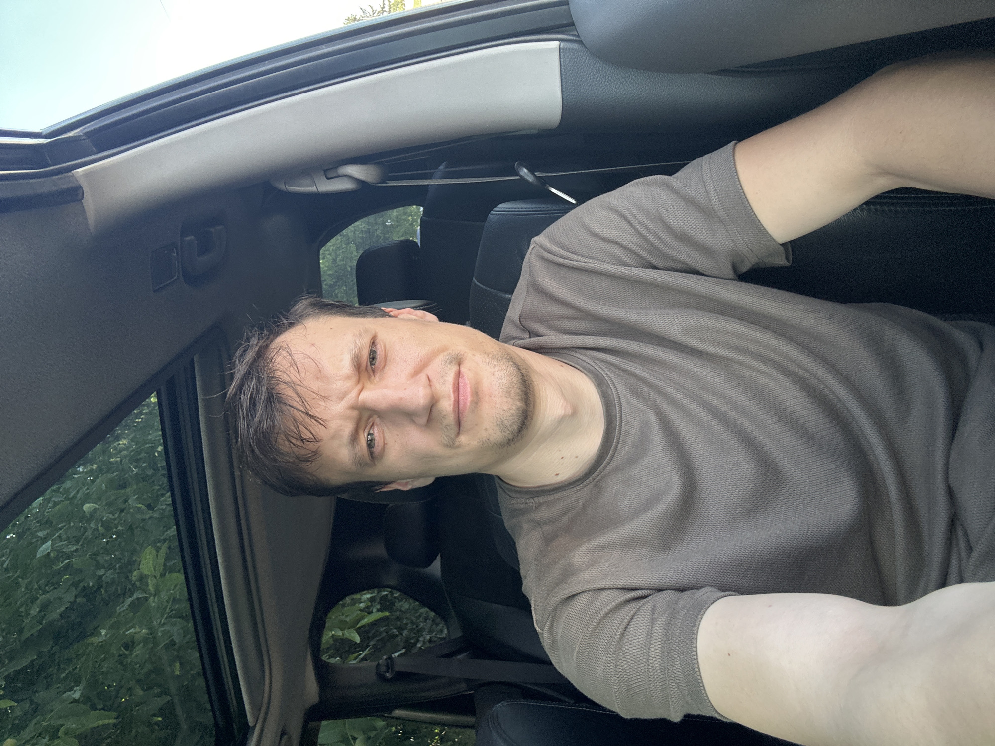 The image shows a man in a car. He is looking at the camera with a neutral expression. The car appears to be parked, with the sunroof open. The interior of the car is visible, including the seats and the dashboard. The man is wearing a grey t-shirt. The background is out of focus, but appears to be a natural environment with trees. The man's face is the central focus of the image. The location of Shatava, Ukraine is not discernable from the image.
