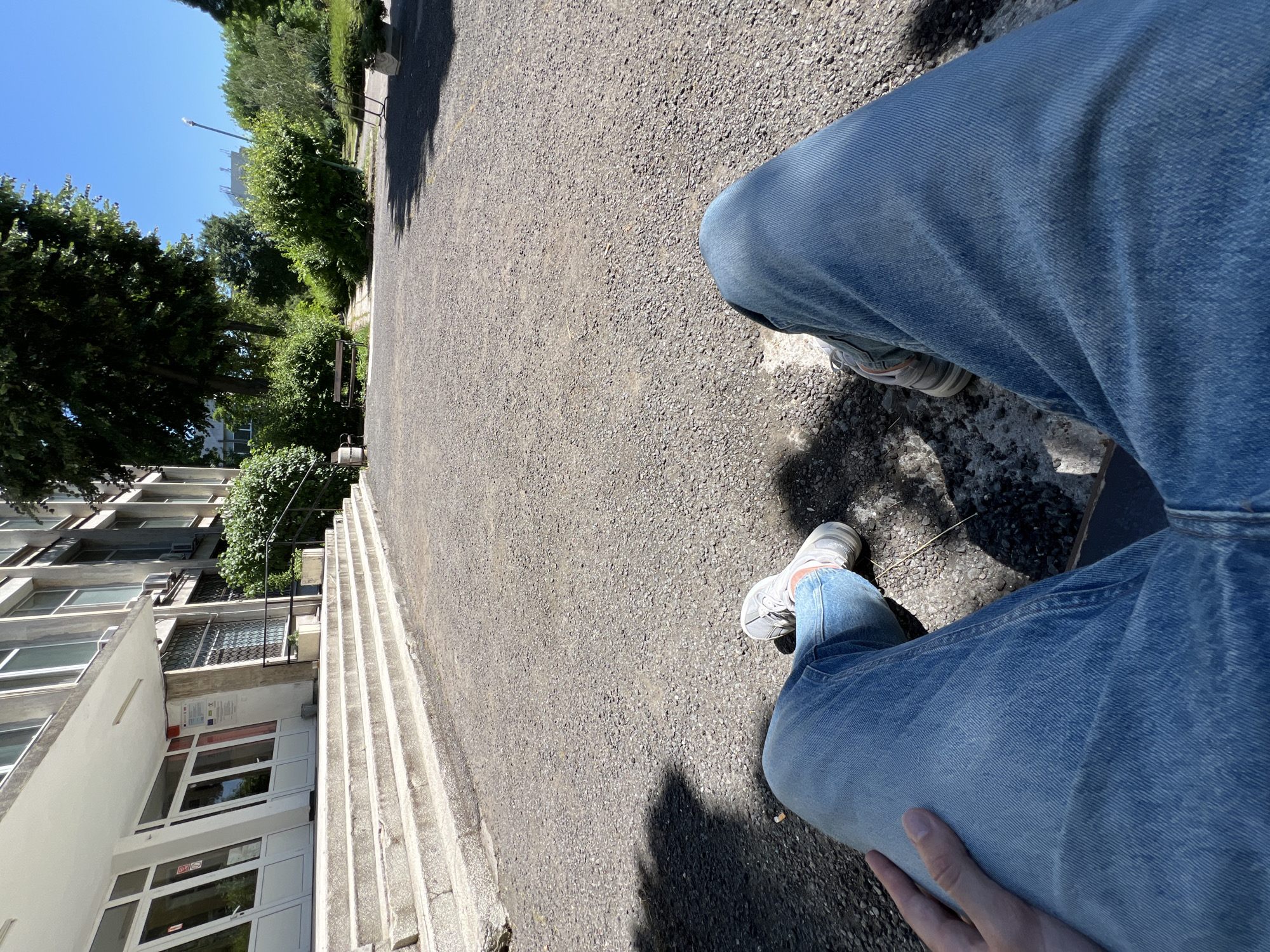 The image depicts a first-person perspective of a person sitting outdoors in front of a building, likely in Burgas, Bulgaria, given the context. The person is wearing blue jeans and light-colored athletic shoes. The foreground is dominated by the person's legs and the gravelly ground. In the background, a multi-story building with numerous windows and an entrance with a wide set of concrete stairs is visible. Adjacent to the building is a lush green area with trees, bushes, and a park bench. The sky is clear and blue, indicating a sunny day. The overall scene suggests a calm, public outdoor space, possibly near an educational institution or administrative building.