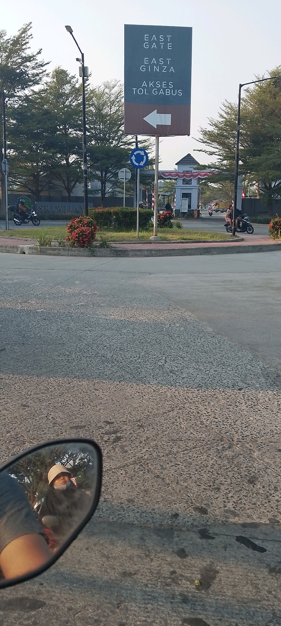 The image shows a street scene in Satriajaya, Indonesia. In the foreground, a motorcycle's rearview mirror reflects a person wearing a light-colored bucket hat and a face mask, sitting on a motorcycle. The background features a large signpost with text that reads "EAST GATE," "EAST GINZA," and "AKSES TOL GABUS" with a white arrow pointing left. Adjacent to the signpost is a roundabout with a blue circular traffic sign depicting arrows. Beyond the roundabout, there is an entrance gate with a tiled roof and columns adorned with red and white flags. Several trees line the street, and other motorcycles and a truck are visible further in the distance. The road surface is asphalt, with some patches of discoloration and debris. The overall lighting suggests it is daytime, possibly late afternoon or early morning given the low angle of the sunlight.