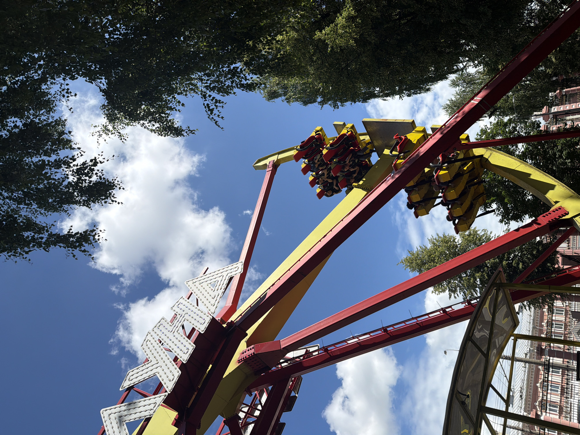 In this eye-level, low-angle shot, a vibrant amusement park ride dominates the frame against a bright blue sky dotted with fluffy white clouds. The ride, a large pendulum structure painted in bold red and yellow, is in motion. Two rows of seats, each a bright yellow with red restraints, are filled with exhilarated passengers, their faces a blur of excitement as they are suspended mid-air.
The ride's massive red and yellow metal framework is intricately detailed with beams and supports, suggesting its complex engineering. A white, triangular structure with small lights is visible on the left side of the ride, possibly part of its decorative elements or warning lights. Lush green trees frame the top of the image, their leaves creating a dappled effect against the sky. In the background, a building with a red roof and white facade, featuring numerous windows, hints at the surrounding amusement park architecture.
The time of day appears to be daytime, likely late morning or early afternoon, given the bright sunlight and clear sky. The weather is pleasant, indicated by the sunshine and scattered clouds. The overall mood is one of exhilaration and fun, typical of an amusement park experience. No discernible text is visible in the image, except for potential branding or markings on the ride that are too small to read. The setting is clearly an amusement park in Izhevsk, Russia, as suggested by the location context.
