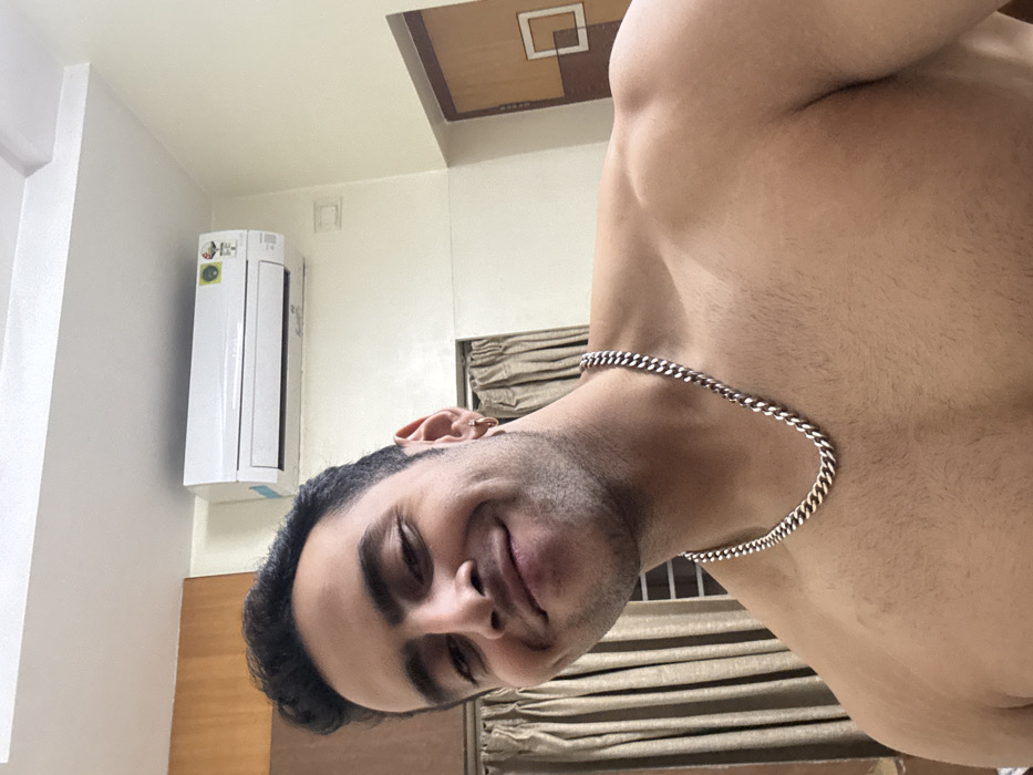 A man with dark hair and stubble smiles into the camera, his head tilted slightly. He wears a silver chain necklace. His bare shoulder and chest are visible. An air conditioning unit hangs on the wall behind him. A beige curtain is pulled shut in the background. The ceiling is light with a geometric design in brown and beige.