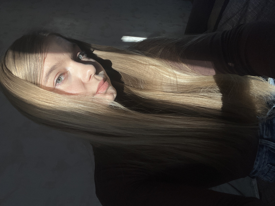 The image depicts a close-up, indoor portrait of a single female subject in the city of Zvezda, Russia. The individual, appearing young, has long, straight, blonde hair that cascades forward, partially obscuring her shoulders and arms. She possesses light-colored eyes, possibly blue or green, and is looking directly towards the viewer. Her skin tone is fair. She wears a dark, long-sleeved top, appearing dark brown or deep red, and light-wash blue denim jeans, visible in the lower left. Strong, directional sunlight illuminates portions of her face, hair, and the adjacent background, creating distinct shadows on the left side of her face and body. A narrow vertical band of direct sunlight is visible on the dark, textured wall behind her on the right. The background is otherwise shadowed, revealing a plain wall and faint outlines of what appears to be a piece of furniture, possibly a bed or sofa, to the right. The subject's pose suggests she is intentionally presenting herself for the photograph, with her head slightly tilted.