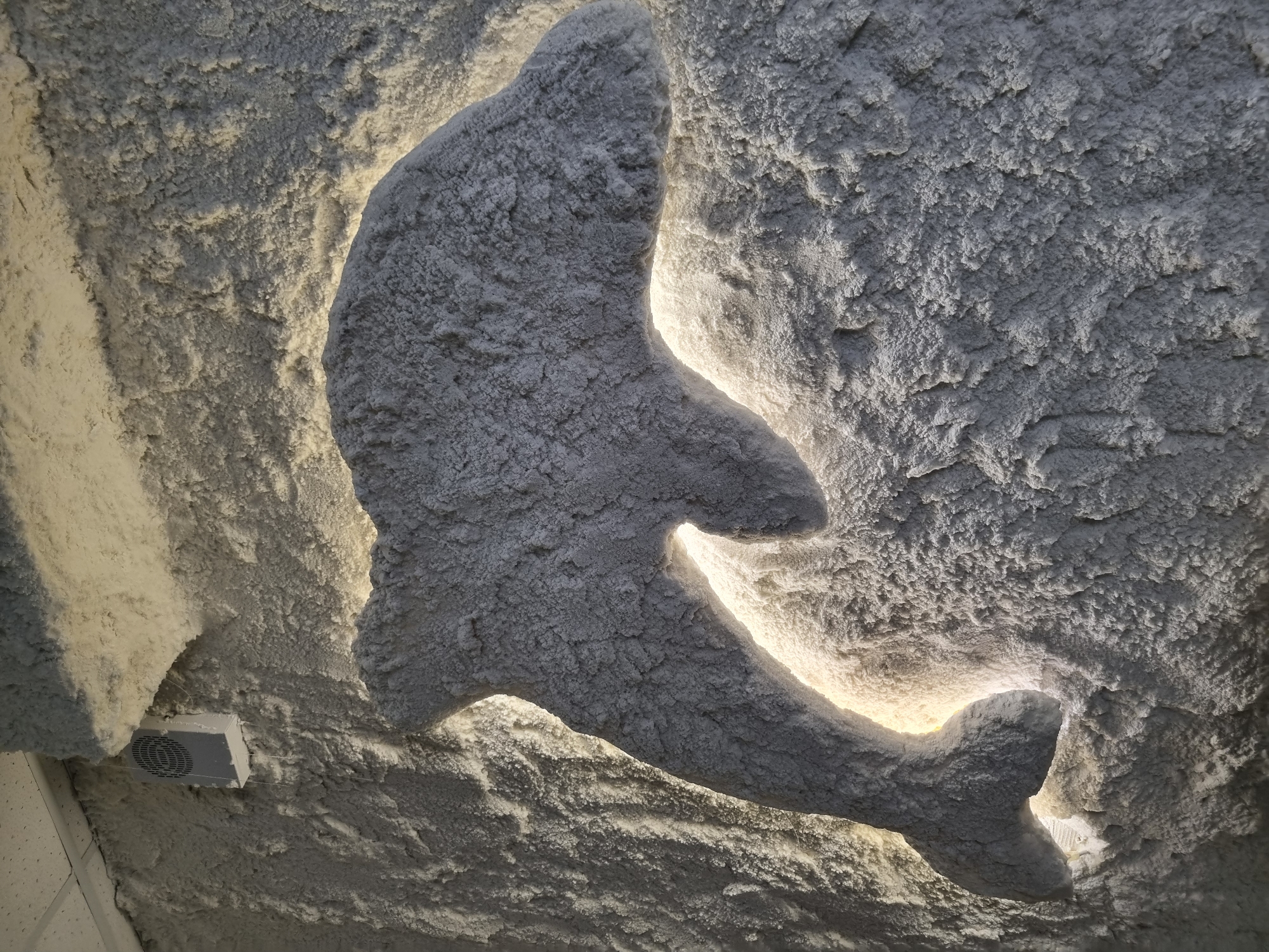A sculpted dolphin made of a light-colored, granular material, possibly salt, is mounted on a textured wall. The dolphin is illuminated from behind, creating a glow around its silhouette. The wall itself is rough and uneven, with visible texture and depth, also appearing to be made of the same granular material. A small white vent with a grill is visible in the upper left corner, attached to a white ceiling. The overall color palette is dominated by shades of white and gray, with warm yellow light emanating from behind the dolphin. This scene is located in the city of Chaikovsky, Russia.