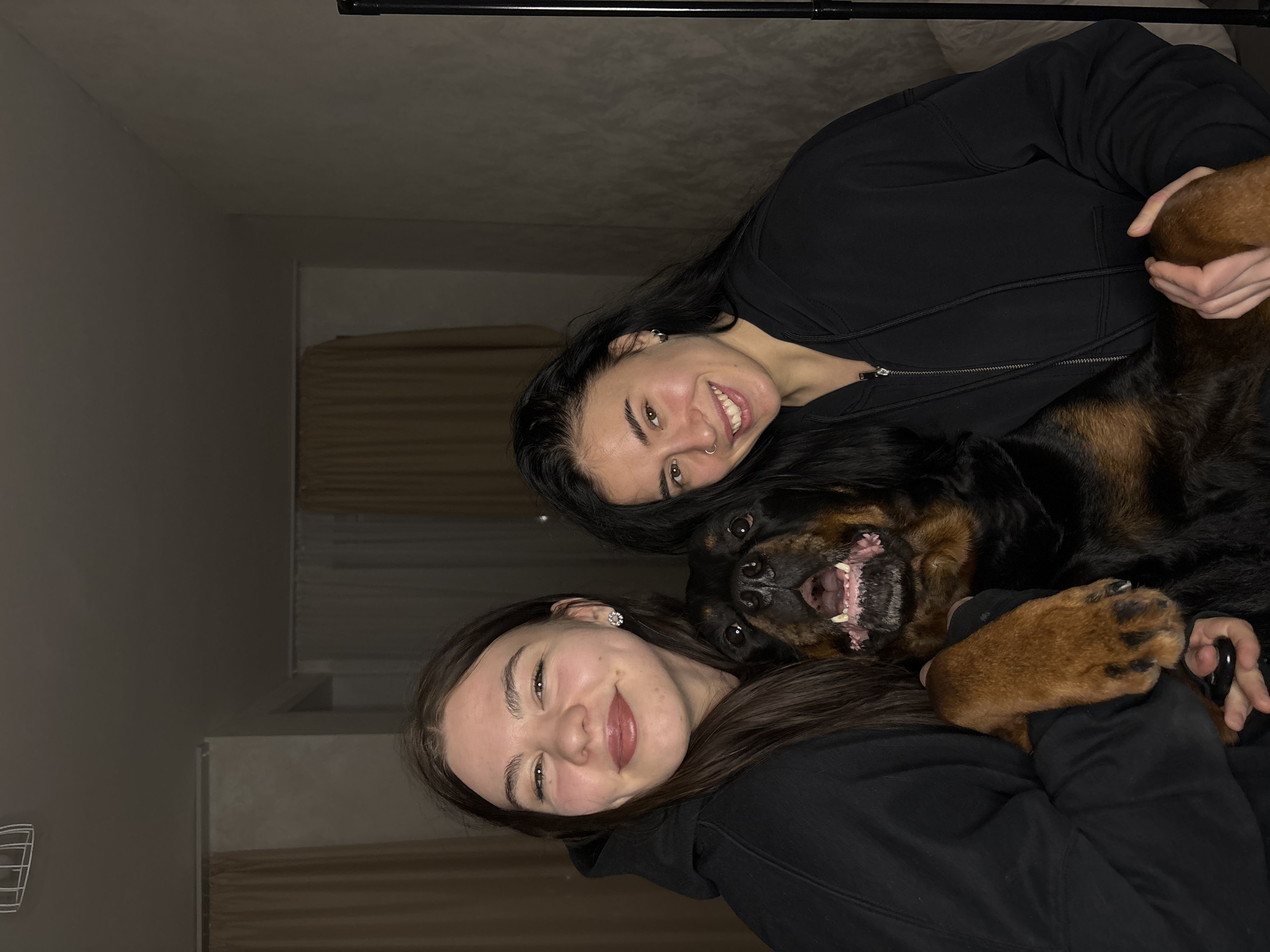This indoor portrait from Perm, Russia, features two young women and a large dog, likely a Rottweiler, arranged closely together. The subjects appear to be lying down or reclining, with the camera positioned slightly above them.
In the foreground, a young woman with long dark hair and a black hoodie smiles genuinely at the camera, showcasing dimples and wearing small, sparkling earrings. Her right arm is gently around the dog, and she is holding one of its large, brown paws. Behind her, another young woman, also with long dark hair and a black hoodie, is visible, smiling broadly. She has a nose ring and her face is positioned above the dog's head.
The dog, a prominent central figure, has black and tan fur and a robust build. It displays a particularly joyful and playful expression, with its mouth open, revealing its teeth and a pink tongue, as if panting happily or "grinning."
The setting is an indoor room with plain, light-colored walls. In the upper background, a dark, horizontal bar or pole is visible. Further in the background, there are hints of a window with horizontal blinds or curtains. The lighting is soft and ambient, suggesting an evening or nighttime shot indoors, contributing to a warm and intimate atmosphere. No visible text is present in the image. The overall emotion conveyed is one of happiness, affection, and companionship between the individuals and their pet.
