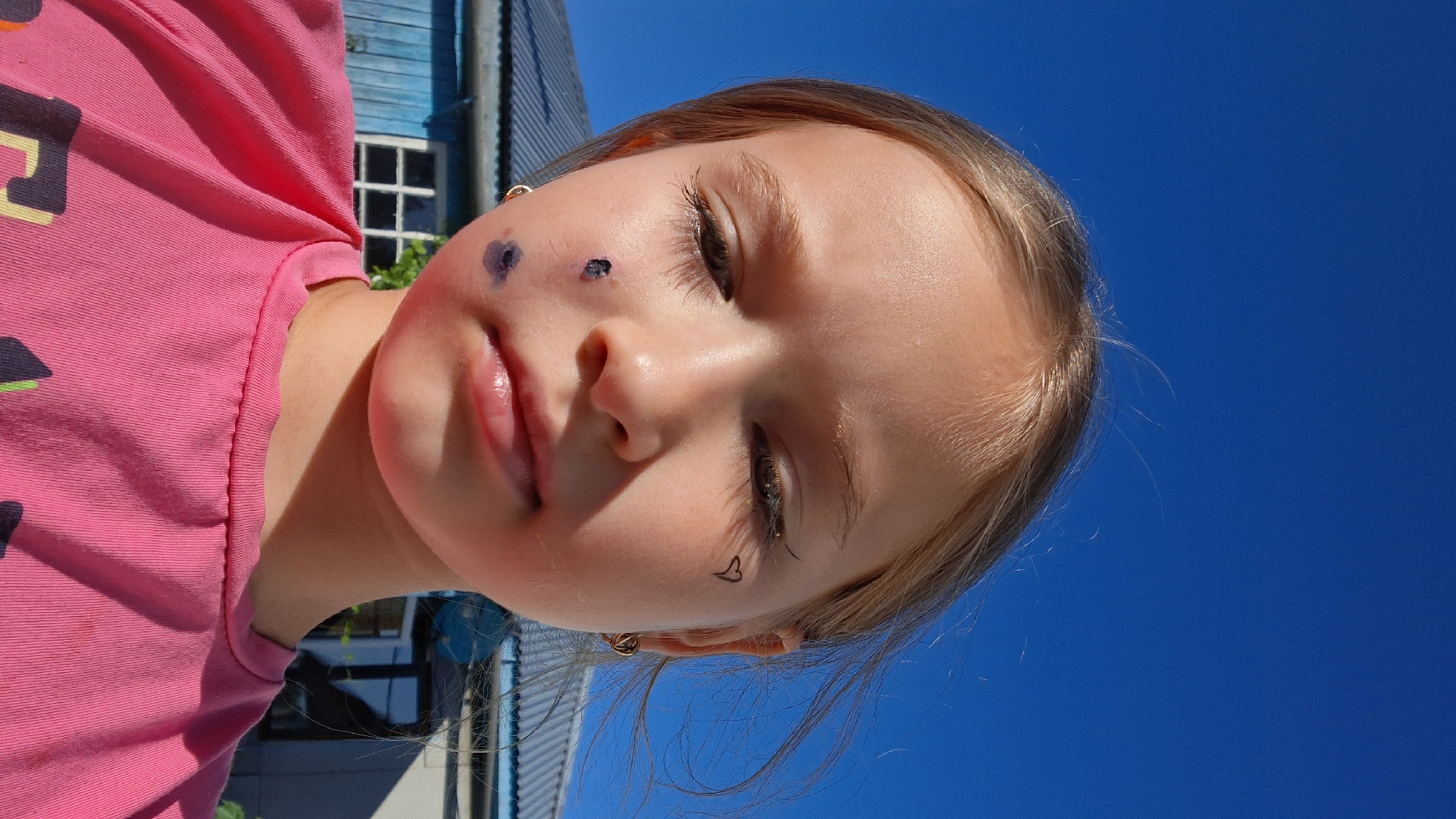 A young girl with blonde hair is looking down, with her face and chest filling the frame. She is wearing a pink t-shirt. There are dark smudges on her left cheek, and a small black heart drawn with eyeliner on her right cheekbone. She also has black eyeliner on her upper eyelids. She is outdoors with a clear blue sky in the background, and the roof of a building, possibly a house, is visible behind her. The lighting suggests it is daytime. The image does not contain any information that would allow for its location to be identified in the city of Semey, Kazakhstan.