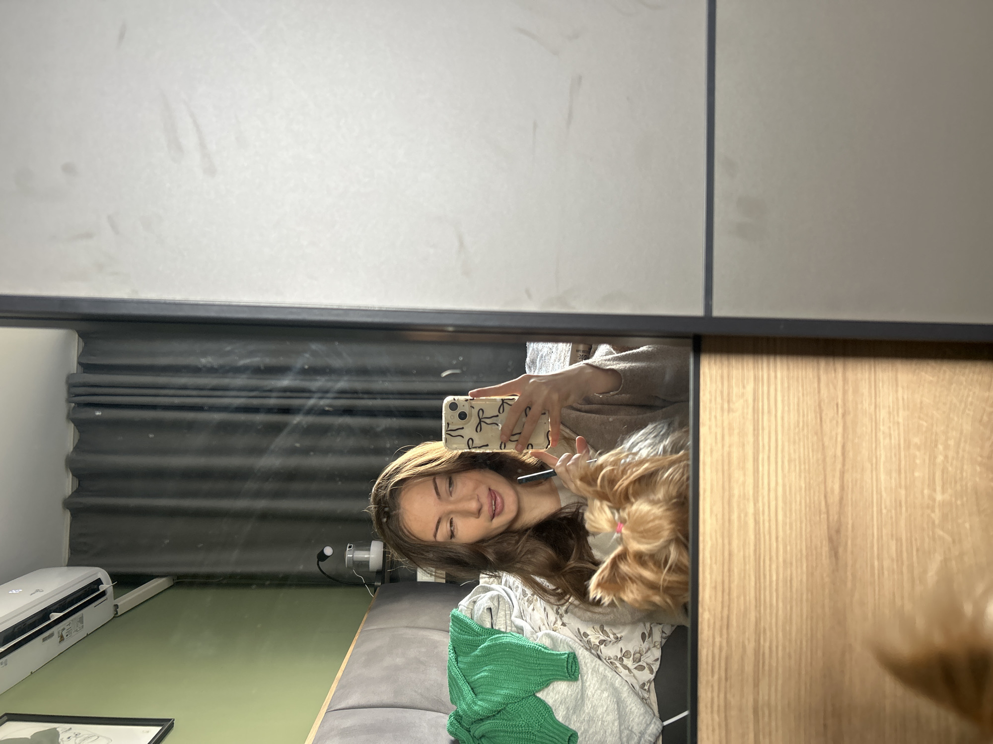 A young woman is taking a selfie in a mirror, likely indoors. She holds a smartphone decorated with bows, and in her other hand is a comb or styling tool. She is smiling and appears to be grooming a small, fluffy, tan-colored dog. The setting includes a gray sofa, a green knit sweater, and a floral-patterned garment. In the background, through the mirror, there is an air conditioning unit, dark window blinds, and green walls. The mirror is framed and appears to be part of a cabinet or wardrobe with light gray and wood-paneled doors. There is nothing in the image that provides a location in Burgas, Bulgaria.