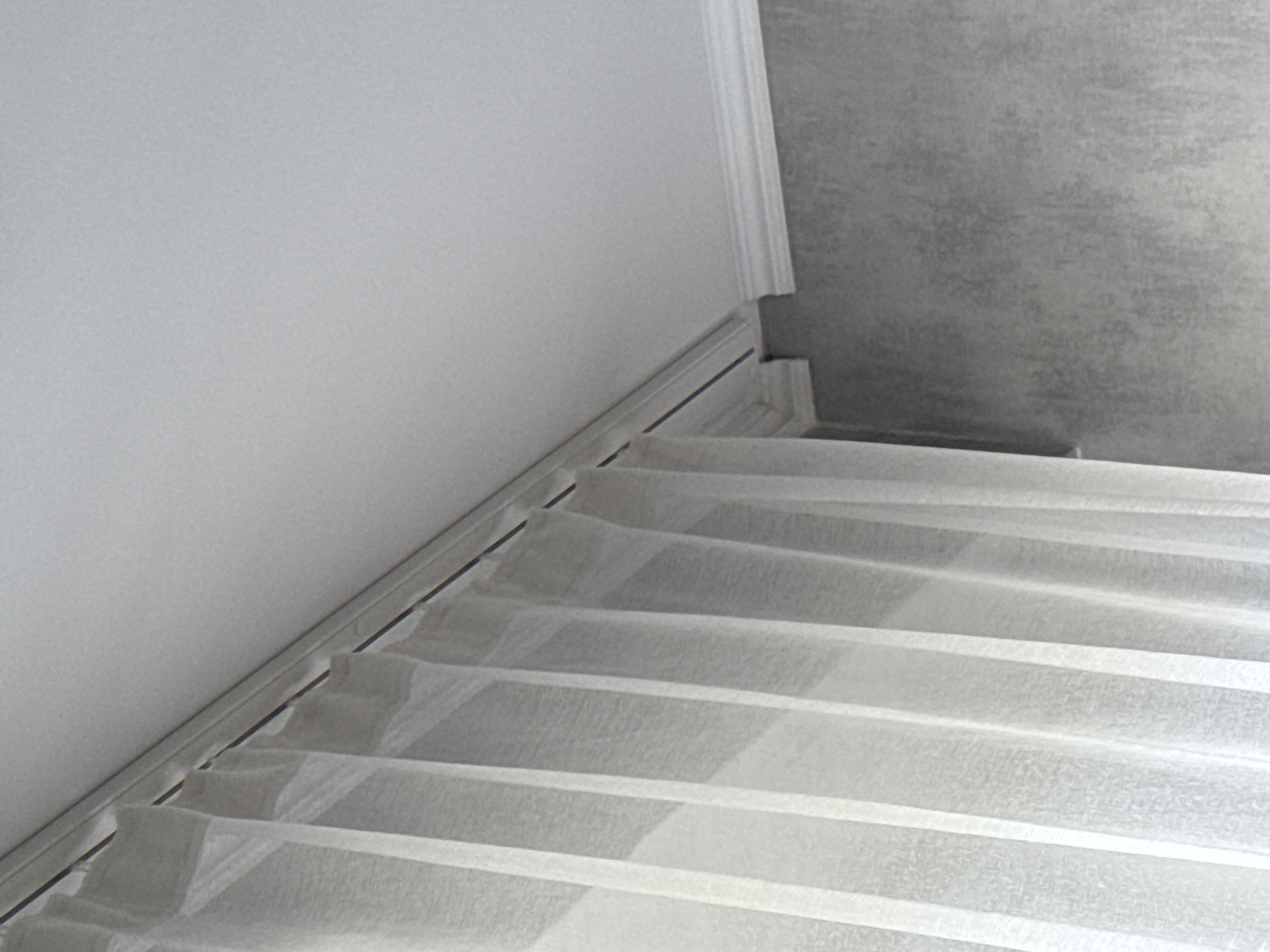 The image shows a downward-facing view of a staircase. The stairs are covered with a white, semi-transparent material, possibly for protection during construction or renovation. The left wall is white and meets the staircase at a baseboard. A decorative crown molding is visible at the top of the wall. The wall on the right side of the staircase appears to have a rougher, gray texture. The location is unknown and cannot be inferred from the image. The city is not identifiable.