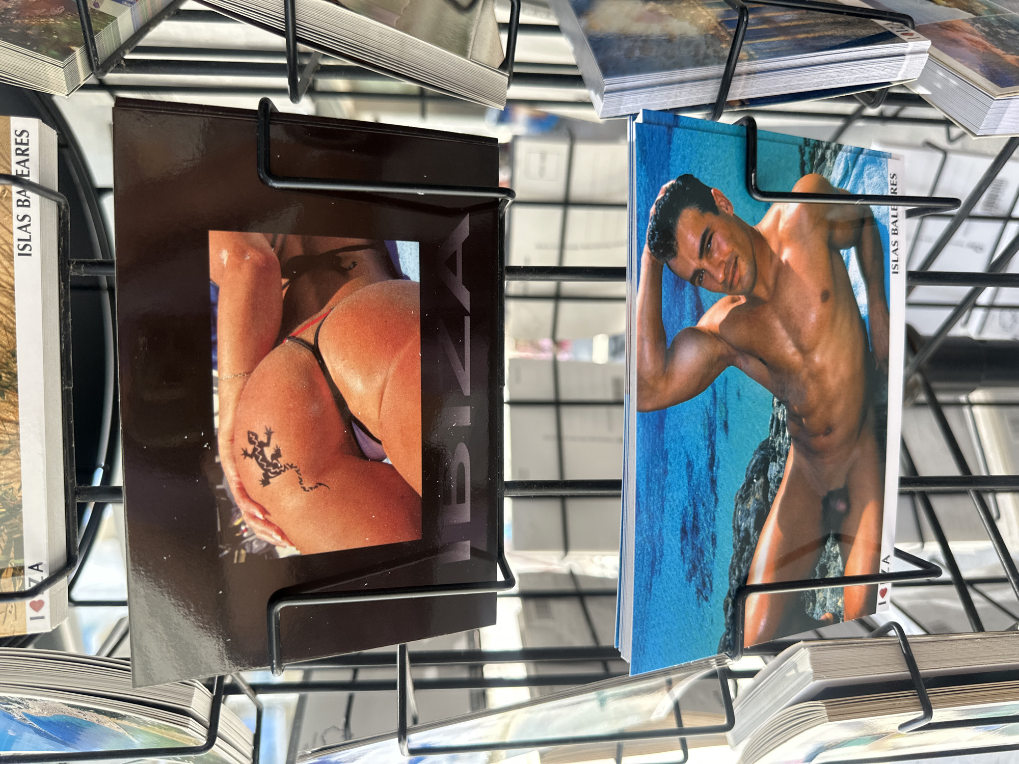 In the image, two postcards are displayed on a metal rack. The postcard on the left depicts a woman's lower back and buttocks with a tattoo of a lizard. The word "IBIZA" is printed next to the image. The postcard on the right depicts a nude male figure. Both postcards appear to be souvenirs from Ibiza, part of the Balearic Islands of Spain. They are held by a black metal wire rack, likely for sale. Other postcards are visible in the surrounding stacks on the rack. The location is presumably a shop or market in Eivissa.