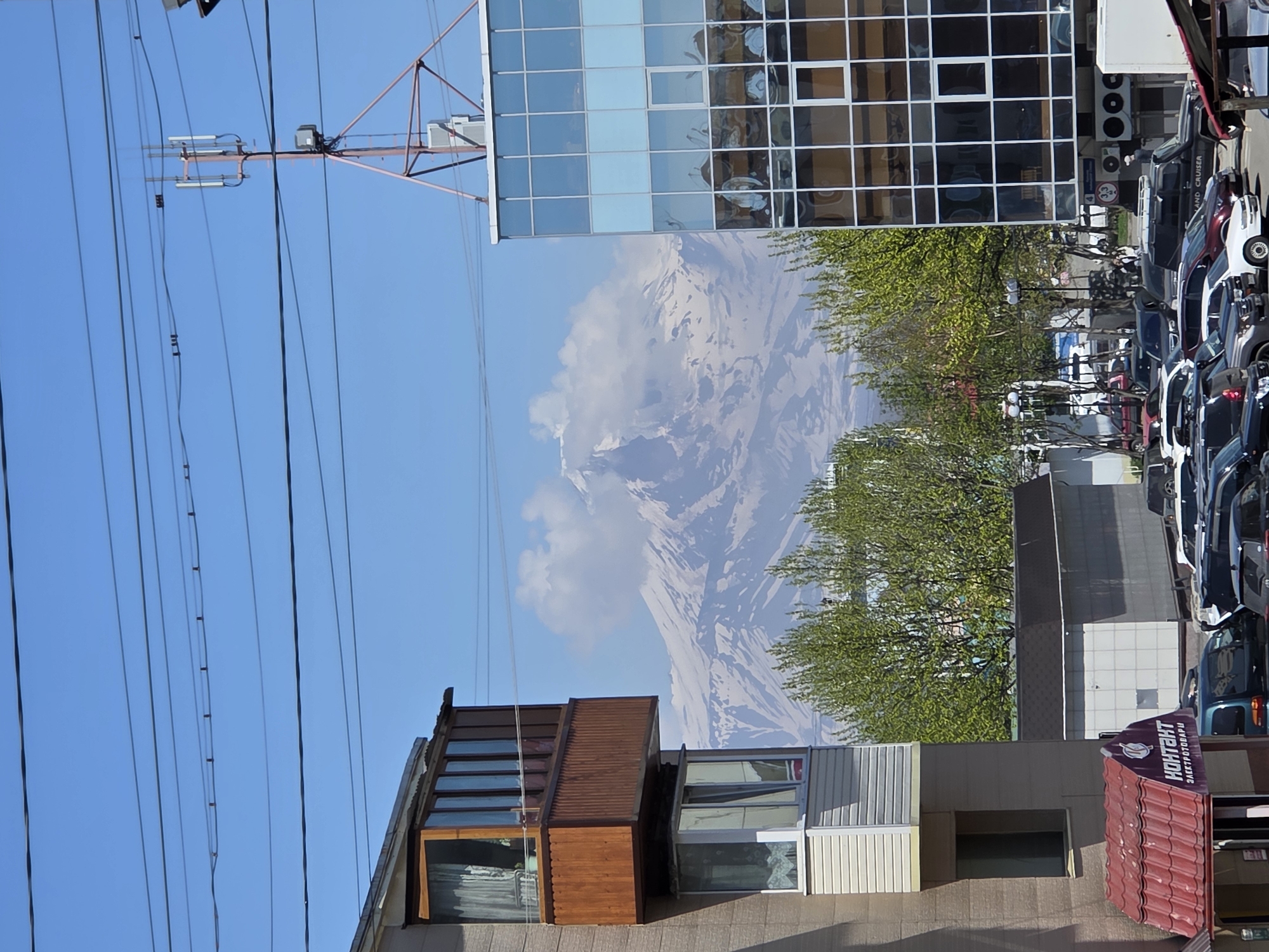 In a city setting under a clear blue sky, a snow-capped mountain looms in the distance, partially obscured by clouds. In the foreground, a modern glass-fronted building stands to the right, reflecting the surrounding environment. To the left, a residential building features a wooden-trimmed balcony and a window. A parking lot filled with various vehicles, including SUVs and sedans, occupies the middle ground. Trees with vibrant green foliage are scattered throughout the scene. Power lines crisscross the sky, and a cell tower is visible against the blue expanse. A small building with a red roof and signage is partially visible in the lower left corner.
