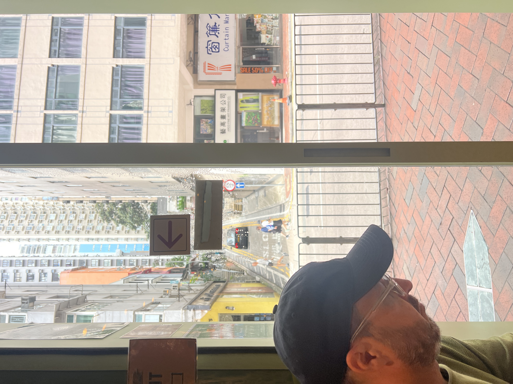 The image captures a view through a glass pane, likely from a cafe or shop, looking out onto a steep, narrow street in Hong Kong. In the foreground, the left side of the image shows the back of a person wearing a black baseball cap and glasses, their face turned down. The glass pane has water droplets on it, creating a slightly distorted view of the scene outside.
The street outside is lined with tall, densely packed apartment buildings of various architectural styles and colors, characteristic of Hong Kong's urban landscape. The street itself slopes steeply upwards, with a black car visible driving up the incline. Two figures, appearing to be pedestrians, are walking down the street. A directional arrow sign with a purple arrow pointing left is affixed to the exterior of the building or on a pole.
Further down the street, commercial storefronts are visible, including one with signage that reads "窗簾" (Curtain) and "Curtain Mar". Another storefront displays artwork. A red fire hydrant is also present on the sidewalk. The street is divided by a metal railing, and beyond that, a patterned brick pavement is visible in the foreground. The overall impression is of a bustling, urban environment with distinct verticality and a sense of being in a tight-knit city.