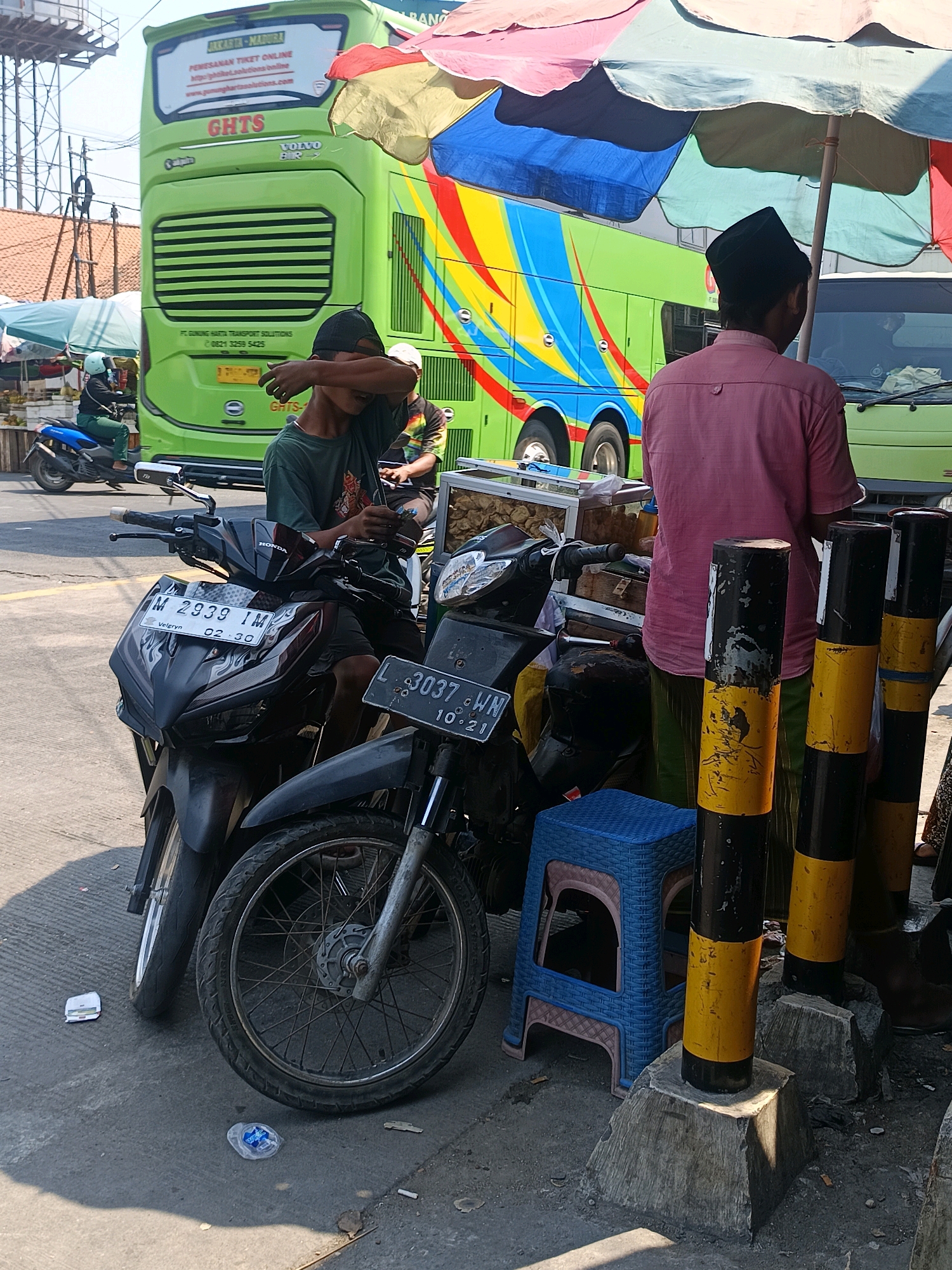 In the city of Blega, Indonesia, a bright green double-decker bus with a colorful, abstract design is parked on a street. The bus has the text "GHTS" and "VOLVO" visible. In the foreground, two motorcycles are parked. The motorcycle on the left has a license plate that reads "M 2939 IM". A young person wearing a dark green shirt and a black cap is seated on this motorcycle, shielding their eyes with their arm. To the right of the young person, a man wearing a pink shirt and a black cap stands near a cart, partially obscured by a large, multi-colored umbrella. A blue plastic stool is placed on the ground next to the motorcycles and the cart. The street appears to be paved with asphalt, and there are concrete bollards with black and yellow stripes nearby. The scene is illuminated by bright sunlight, casting shadows on the ground.