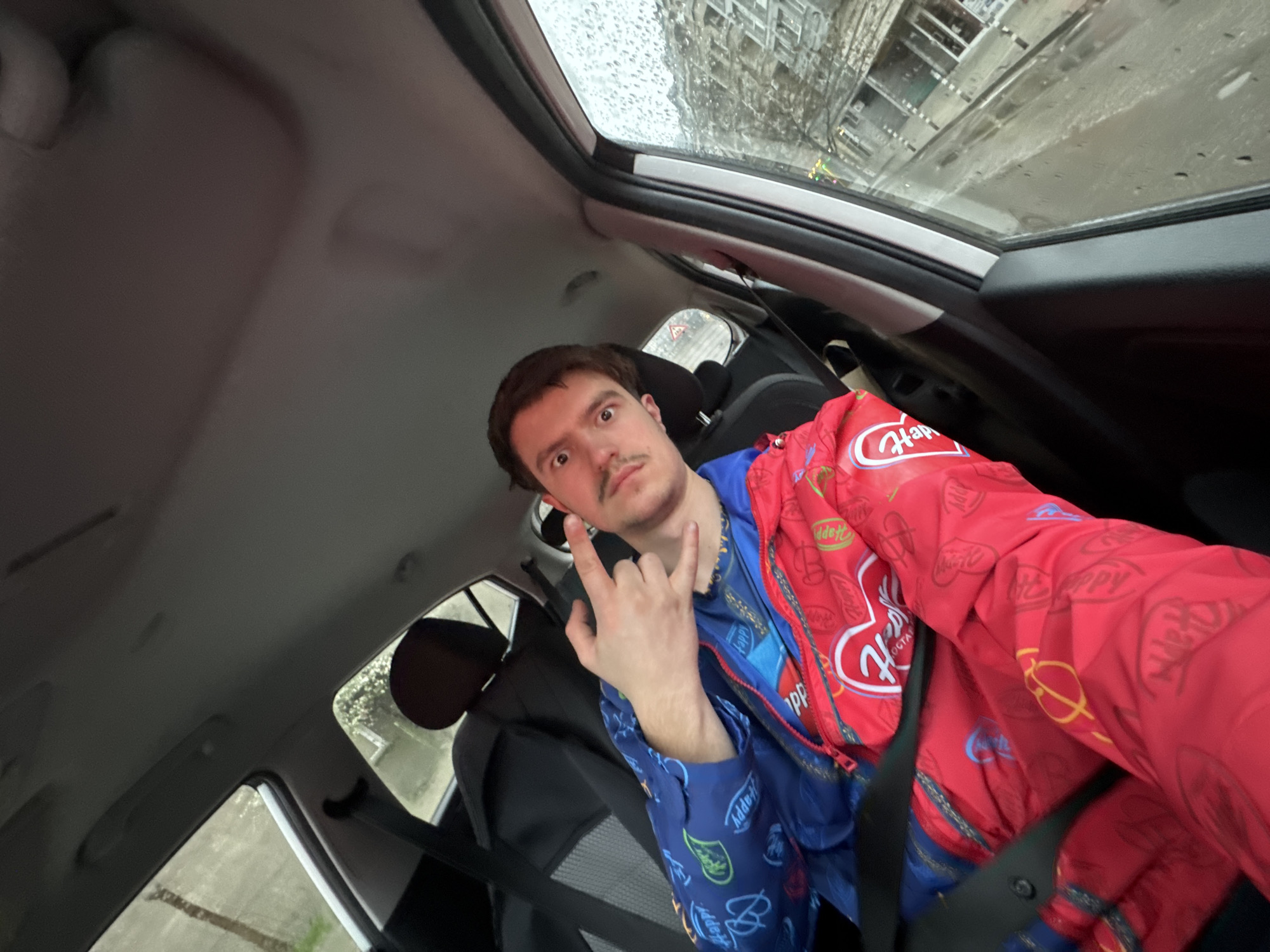 A man is sitting in the driver's seat of a car, likely in Sofia, Bulgaria, based on the urban environment visible through the windshield, which shows buildings typical of the city. The car has a sunroof, which is open and lets in some of the ambient light. The man, fair-skinned with brown hair and a mustache, is wearing a split-color jacket; one side is blue with graphics, while the other is red with "Happy" logos and graphics. He's making a "rock on" hand gesture. The car's interior includes a black dashboard and black seats. It appears to be raining outside. The perspective is from an slightly elevated angle, looking down into the car.