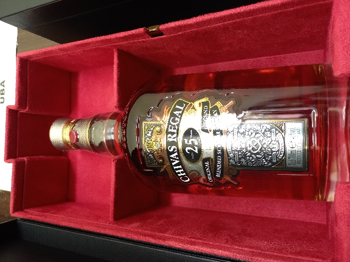 A bottle of Chivas Regal 25 Year Old Blended Scotch Whisky is prominently displayed within an open, red velvet-lined presentation box. The clear glass bottle contains an amber-colored liquid and features a gold-toned, ribbed screw cap with a small red circular seal. The main label reads "CHIVAS REGAL 25 Years ORIGINAL BLENDED SCOTCH WHISKY," with a lower label detailing "70cl / 700ml e" and "40% vol.," indicating production in Scotland. The interior of the box is molded to cradle the bottle, while its exterior, partially visible, is black. A white object displaying the letters "UBA" is visible in the upper background. The scene is located in the city of Oturkpo, Nigeria.