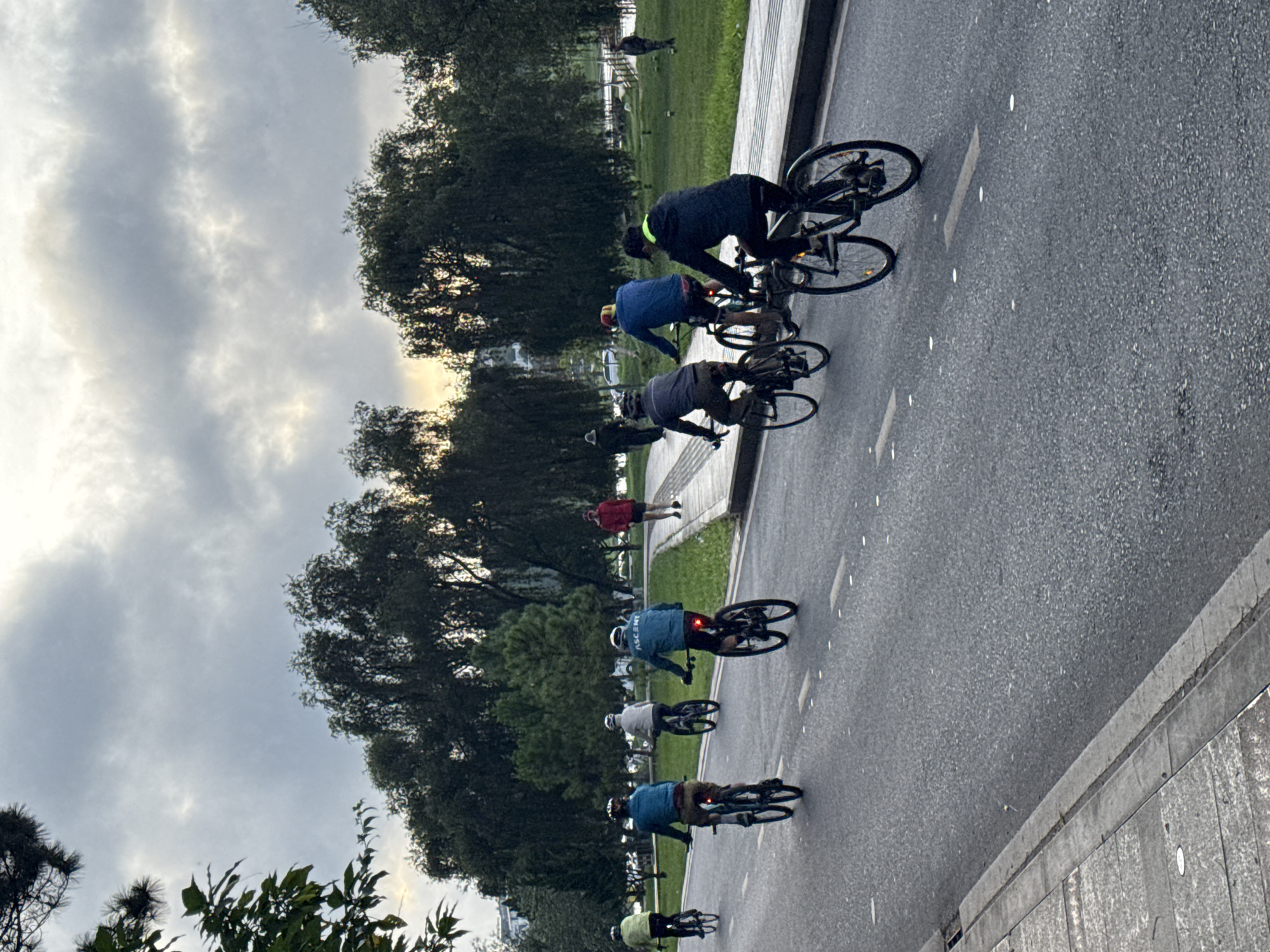 The image captures an outdoor scene in Phường Xuân Hương - Đà Lạt, Vietnam, showing five individuals cycling away from the camera on a paved asphalt road, which features white dotted lane markings. The sky is overcast with light, textured clouds, but a band of soft, warm light is visible at the horizon, indicating either dawn or dusk. From left to right on the road, the cyclists are dressed in a yellow-green top, a blue jacket, a light beige jacket, a blue jacket with "ASCENT" emblazoned on the back, and a dark jacket with yellow trim. All are riding dark-colored bicycles, with several exhibiting active rear brake lights. A concrete sidewalk runs parallel to the road on the right, bordered by a wide, well-maintained grassy expanse populated by numerous tall, dense trees. Three additional individuals are present in the background: one person in a red jacket walks along the sidewalk, while two others in darker attire are situated further back within the treed area. The distant background reveals a dense line of trees and a subtle hint of a body of water, consistent with the location of Hồ Xuân Hương.
