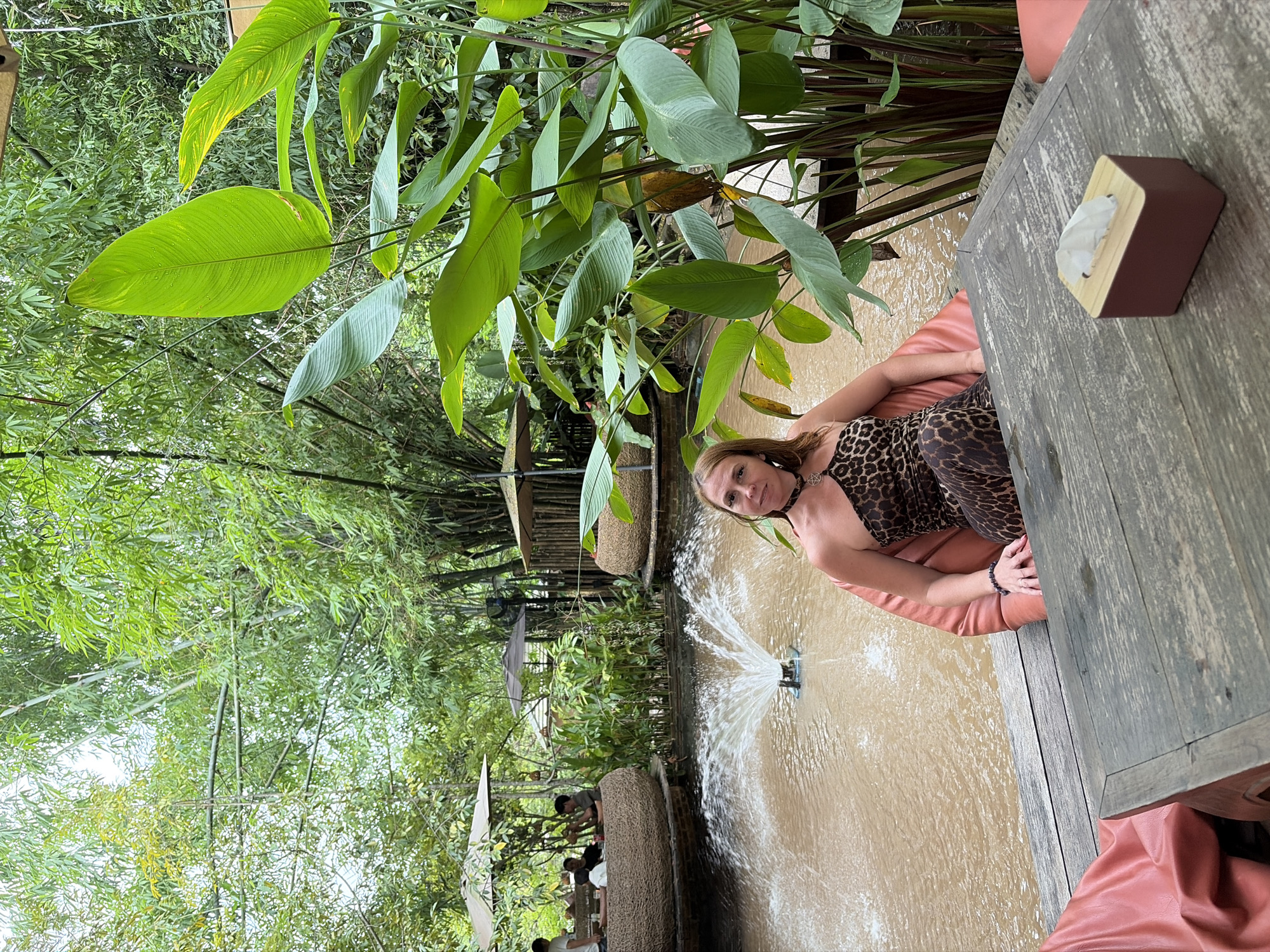 A woman is sitting on a large, orange beanbag chair next to a wooden table. She is wearing a leopard-print, one-shoulder dress and a necklace. In front of her is a shallow pool of water with a small fountain. The area appears to be an outdoor seating area for a restaurant or cafe, with more seating and umbrellas visible in the background. Lush green vegetation, including bamboo and large-leafed plants, surrounds the scene. The image was taken in Tambon Mae Raem, Thailand.