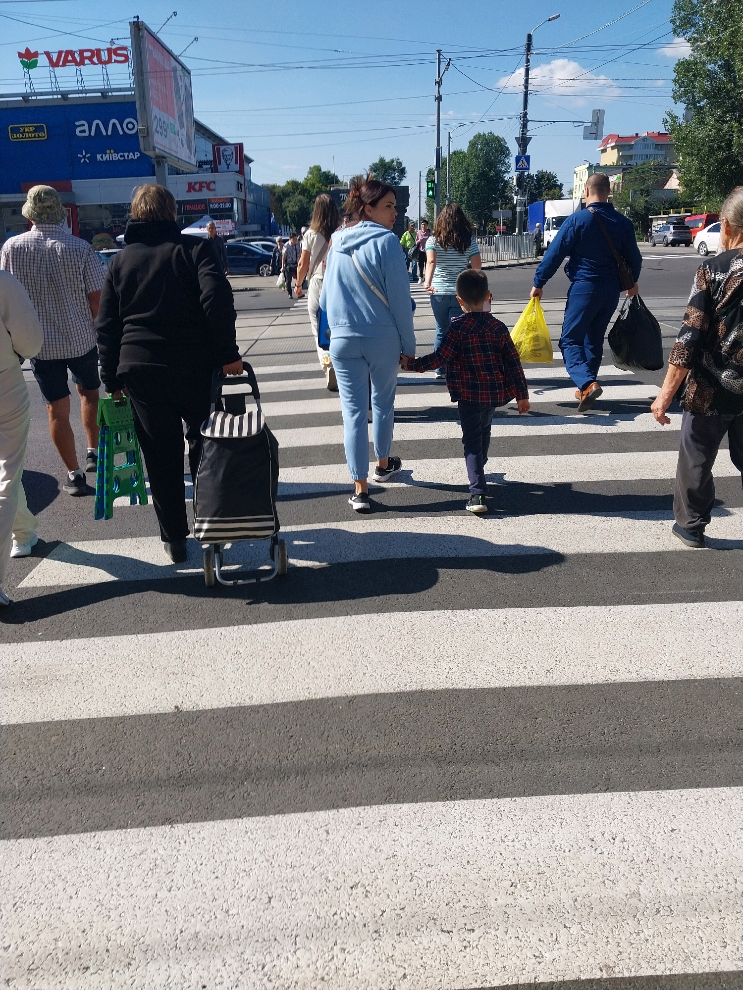 The image shows a sunny day in Dnipro, Ukraine, with several people crossing a pedestrian zebra crossing. In the background, a building with "VARUS" and "allo" signs is visible, suggesting this is a commercial area. One person is pulling a shopping trolley, another is carrying a green folding step stool, and another person is holding two plastic bags. A child is holding hands with an adult as they cross the street. Cars are also visible in the background. The sky is clear and blue, with some clouds. The overall scene depicts a typical urban environment with everyday activities.