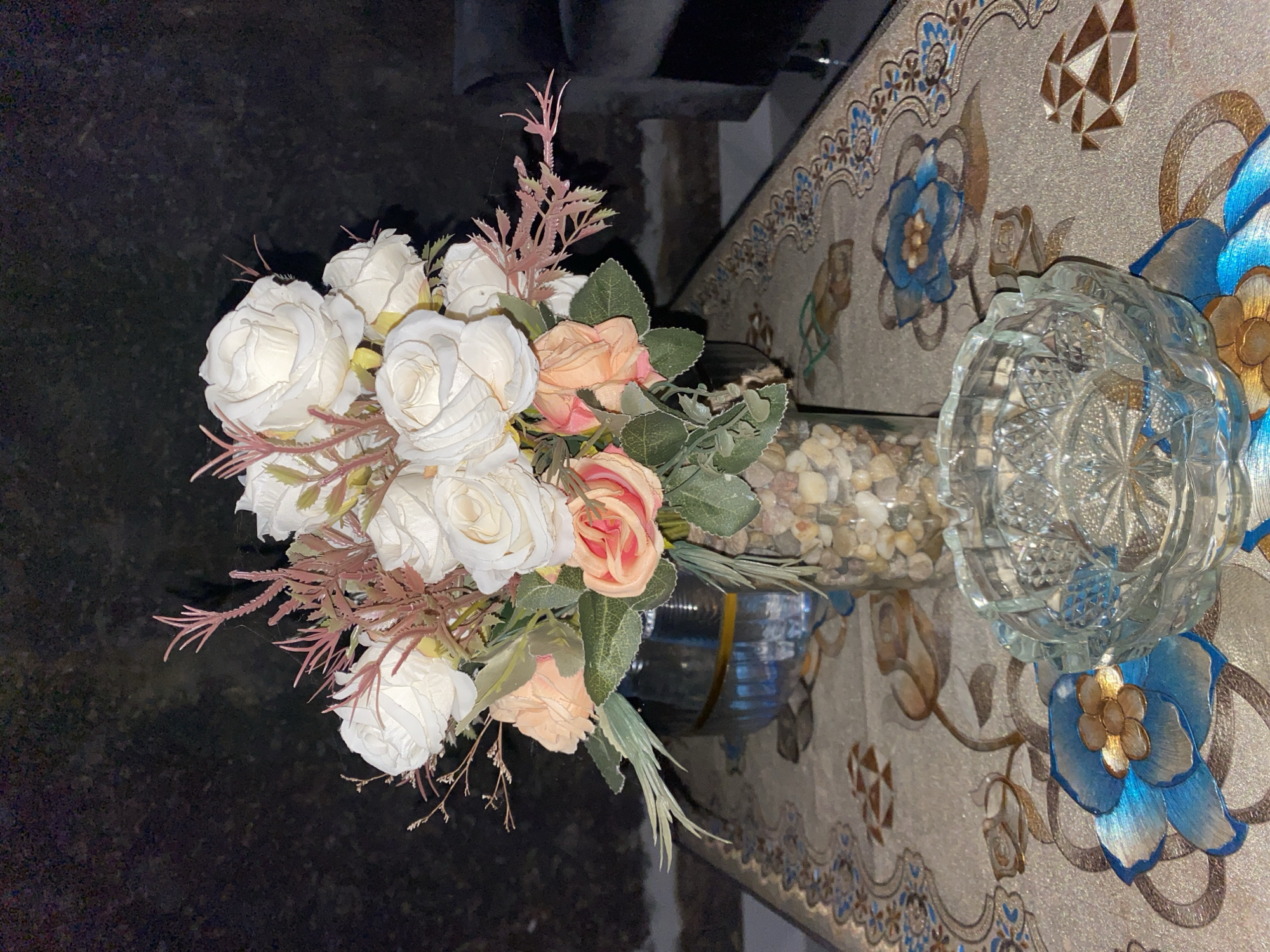 A still life photograph displays a bouquet of artificial roses, predominantly white with some peach and pink hues, arranged in a glass vase filled with small stones. The vase sits on a patterned tablecloth featuring floral motifs in blue, gold, and brown. In the foreground, an empty, faceted glass ashtray rests on the table. The background is dark and out of focus, suggesting an indoor setting. The image does not provide any visual information to place it in Bontoharu, Indonesia.