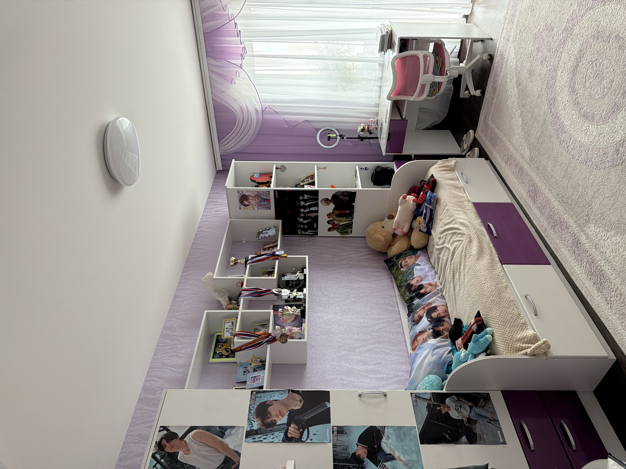 The image depicts a child's bedroom. The walls are primarily white and purple. A bed with white and purple storage drawers along the side is adorned with plush toys and a pillow featuring portraits of people. A white desk with a pink chair sits near a window with light curtains. Bookshelves display trophies, memorabilia, and pictures, some of which appear to be of popular culture figures. The floor is covered with a purple patterned rug. Based on the cultural artifacts and context clues, this scene is most likely located in Penza, Russia.