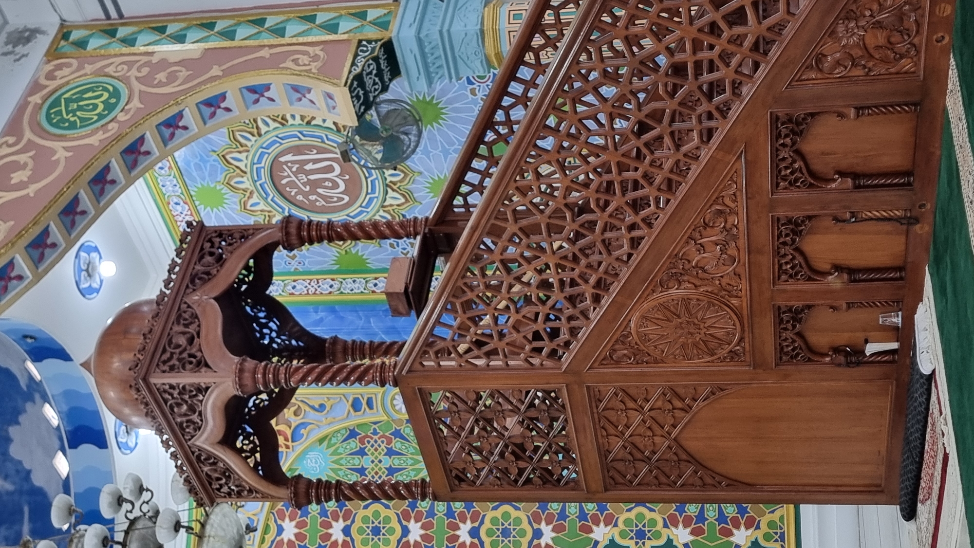 This image shows a wooden minbar, a raised pulpit used in mosques for religious sermons, located in the city of Keudekruenggeukueh, Indonesia. The minbar is intricately carved with geometric patterns and Islamic calligraphy, and it has a domed roof. The background features colorful mosaic tiles with Arabic inscriptions and a fan mounted on the wall. The floor is covered with prayer rugs. The scene appears to be inside a mosque, with chandeliers visible on the ceiling. The image is taken from a low angle, looking up at the minbar. There are no people visible in the image.