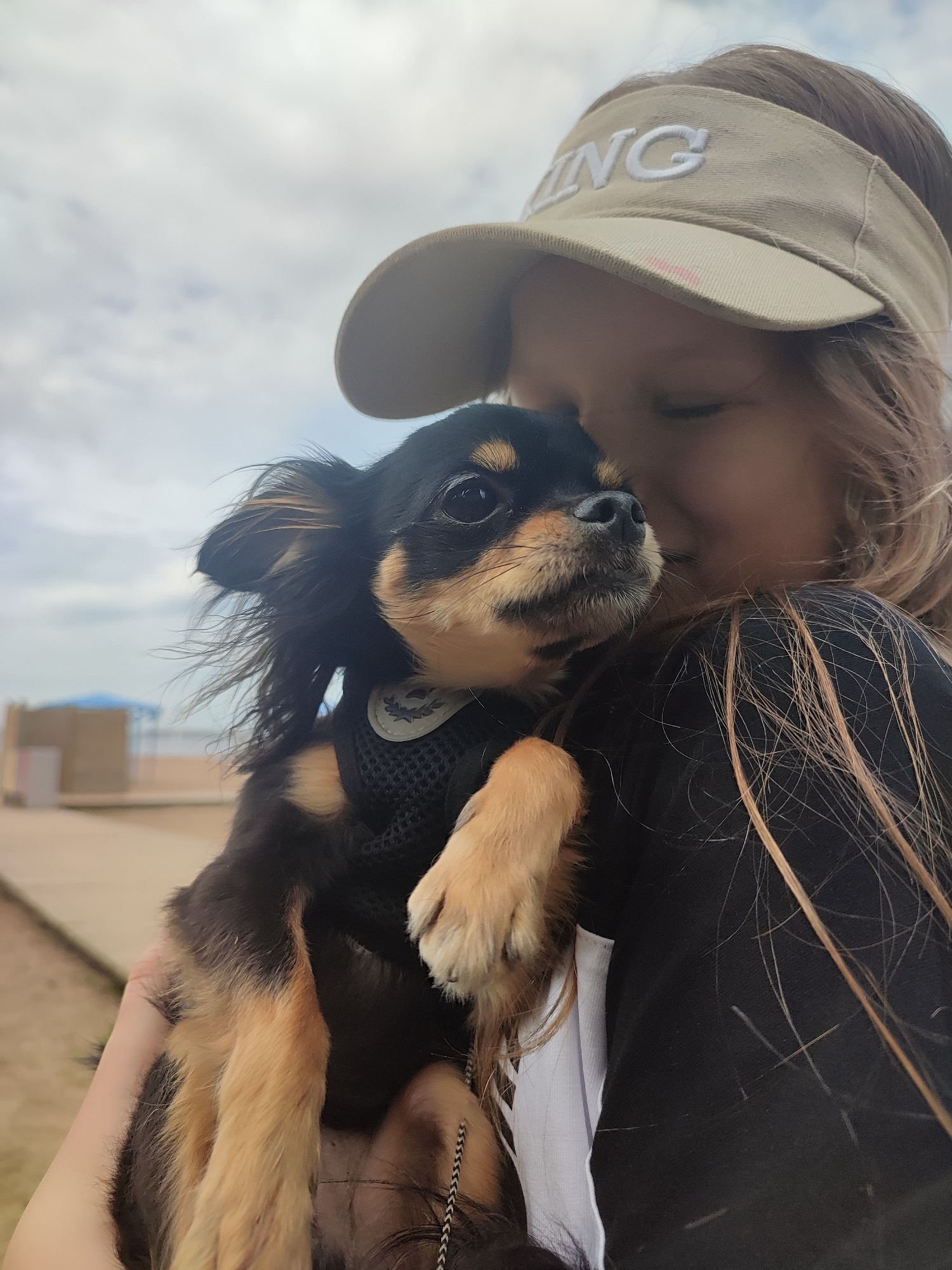 A young fair-skinned female is holding a black and tan chihuahua dog against her body. The dog has a black harness on. The female has her eyes closed and is wearing a beige visor with lettering on the front. The location appears to be on a beach with a structure with a blue roof visible in the background. The sky is overcast.