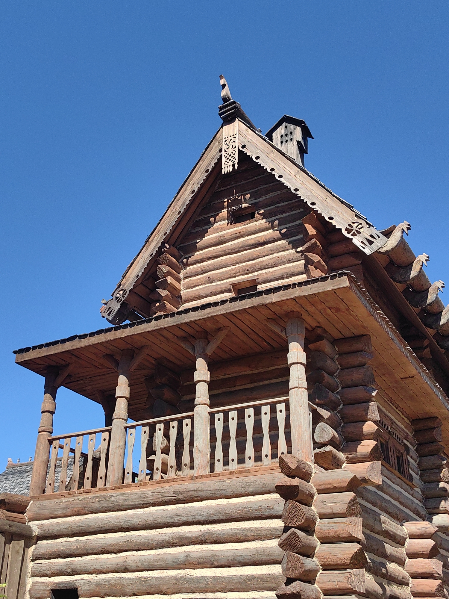 The image shows a detailed, close-up view of a traditional log cabin, characteristic of Russian architecture, against a clear blue sky. The cabin is constructed from dark brown, rough-hewn logs with visible chinking between them. It features a steeply pitched roof with decorative wooden carvings along the eaves, including what appear to be stylized bird figures and intricate patterns. A prominent covered porch with wooden railings and sturdy support posts extends from the upper level of the cabin. The log construction is evident throughout the visible portion of the building, with interlocking corner joinery clearly visible. The bright sunlight casts shadows, highlighting the texture of the wood and the details of the craftsmanship. No people are visible in the image. The specific location is identified as Терехово-2 Снт, Russia.