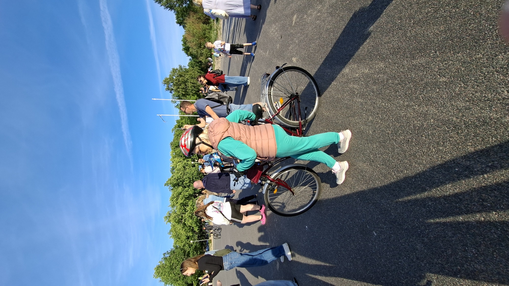 In Tallinn, Estonia, on a sunny day with a clear blue sky and sparse clouds, a group of people are gathered on a paved area, likely a promenade or park, near a treeline. The foreground is dominated by a woman wearing a red and silver helmet, a light pink puffer vest over a turquoise tracksuit, and grey sneakers. She is standing next to a red bicycle, holding its handlebars. Her gaze is directed to her left, and she appears to be looking at her phone, which is held up with her right hand. Behind her, other individuals are visible, some walking, others standing. A young boy in a white t-shirt and black shorts is standing to the right of the frame, facing left. The overall impression is of a casual outdoor gathering.