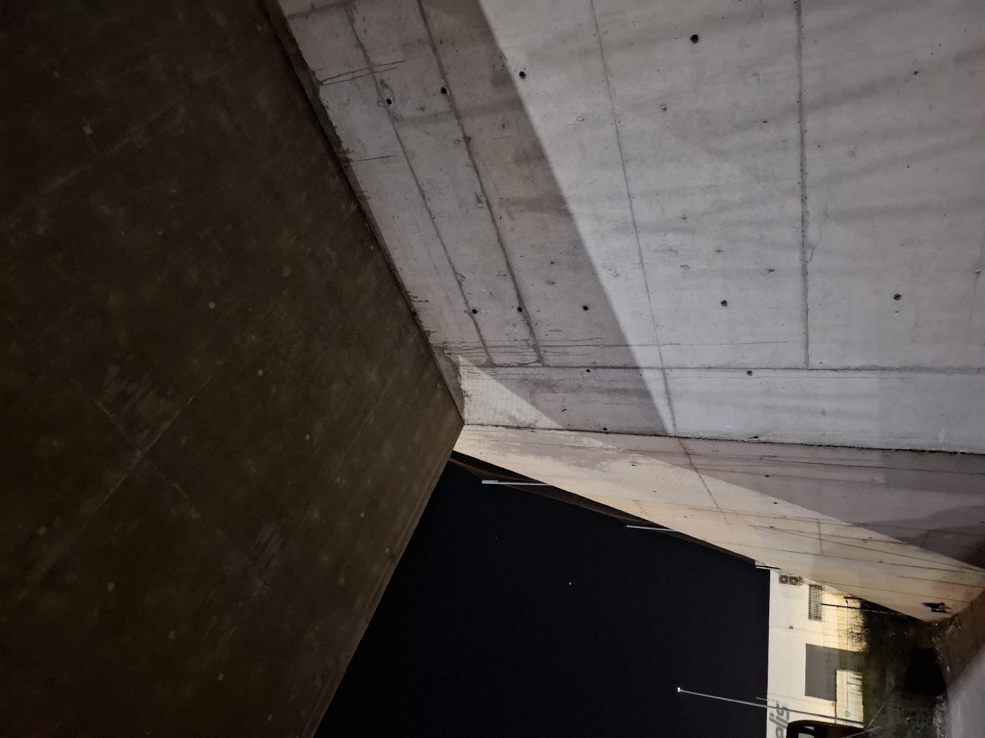 The image depicts the underside of a concrete overpass or bridge structure, likely at night. The location is identified as Quart de Poblet, Spain. Two concrete walls meet at an angle, framing a view of the night sky and a portion of a building and street. The concrete shows signs of wear and construction details such as bolt holes and seam lines. The lighting is dim, illuminating parts of the concrete structure and the nearby building. The building appears to be a low-rise commercial structure with windows and signage.