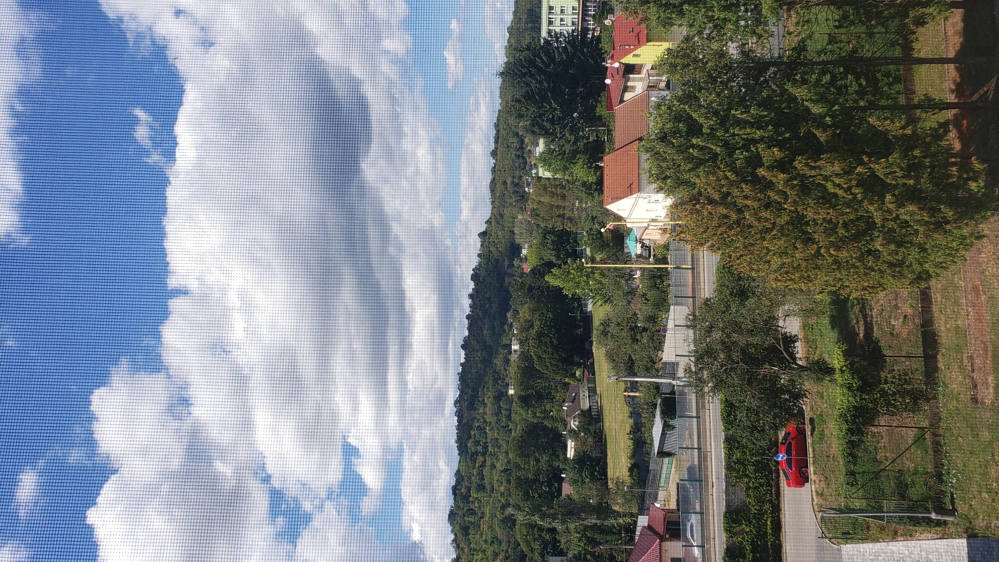 This is an outdoor, medium-distance, vertical shot of a suburban landscape viewed through a window screen. The sky is bright blue with scattered white clouds. A tree-covered hill serves as the backdrop for a cluster of residential buildings with red and yellow tile roofs. In the foreground, a red car is parked on the side of a road, with a fence and lush green vegetation bordering the scene.