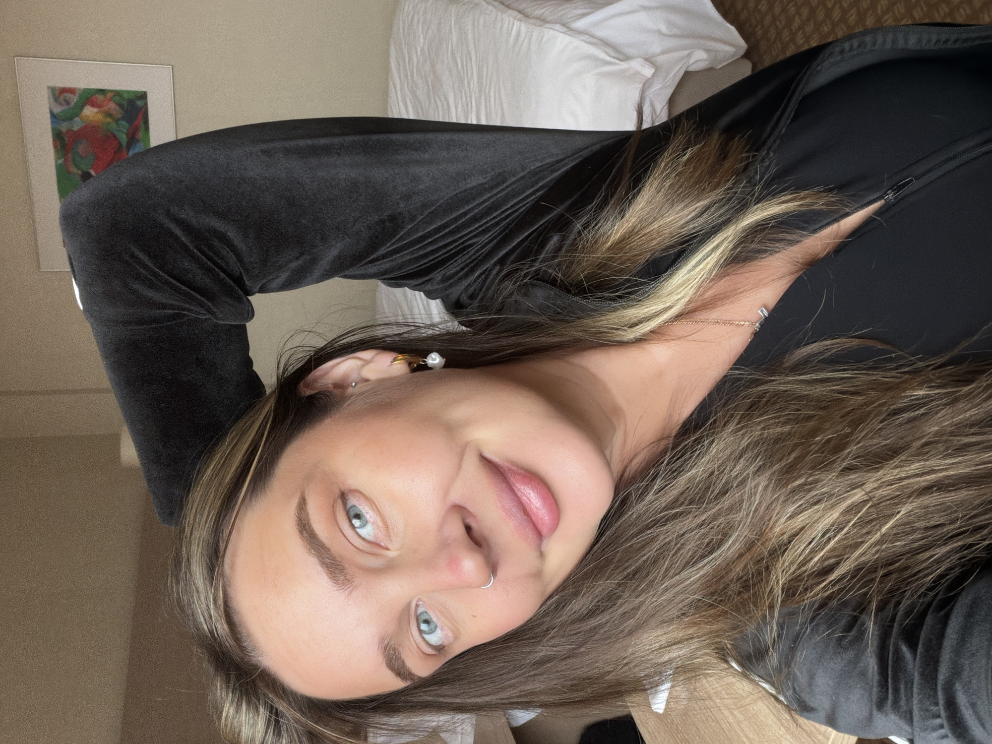 A woman with blue eyes and brown and blonde highlighted hair is looking at the camera. She is wearing a black, long-sleeved top. She has a pearl earring in her left ear and a nose ring in her left nostril. Behind her is a wall with a framed picture and a white bed. The setting is a room, likely a hotel. There is no evidence in the image to suggest this image was taken in Moscow, Russia.
