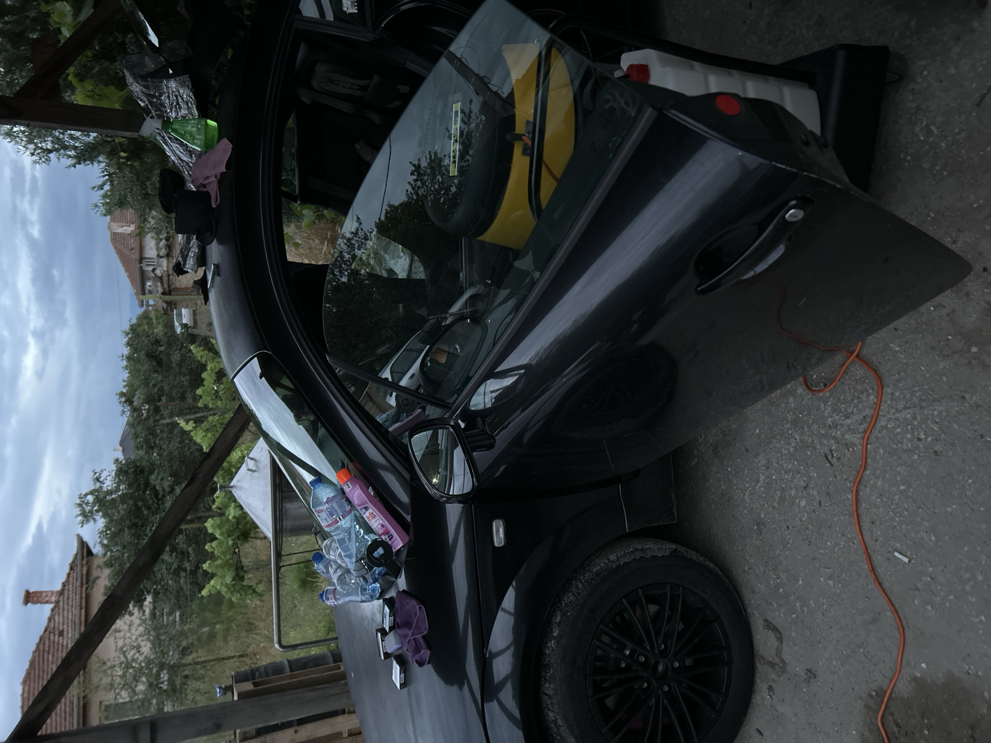 The image depicts a black Audi TT parked outdoors in Benkovski, Bulgaria, under a cloudy sky. The driver's side door is ajar, revealing the car's interior. On the hood of the car, several plastic bottles, a purple microfiber cloth, and packs of cigarettes are visible. A green spray bottle and a pink cloth are placed on the roof of the car. Inside the car, a yellow object, possibly a tool bag or a speaker, is visible. A white plastic container with a red cap is located next to the car, on the right side. An orange electrical cord lies on the ground in front of the car, with a cigarette butt near it. In the background, a house with a red tile roof and trees can be seen. The setting appears to be a yard or a garage area.