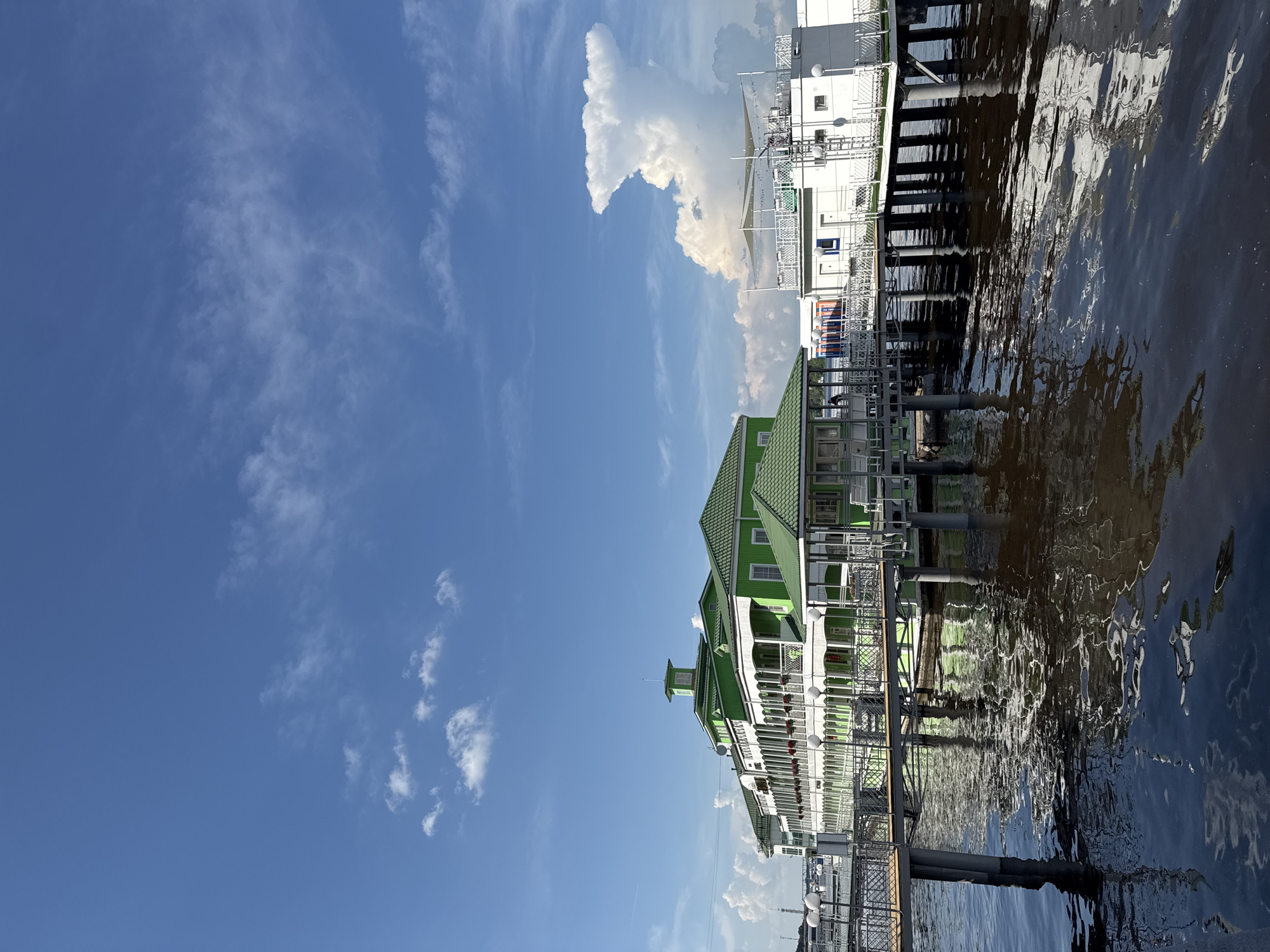 This image captures a waterfront scene in the city of Zavolzhye, Russia. In the foreground, dark, rippling water reflects the sky and the structures on the shore. A metal pier with dark railings extends from the left side of the frame, supported by gray pilings. On the pier, a long, multi-story building painted in bright green and white stands prominently. It features a green roof, white balconies with ornate railings, and numerous windows. The building is elevated and appears to be situated on the water. To the right of this green building, a white, multi-level structure with a flat roof and a large, white, mushroom-shaped cloud formation in the sky behind it. The sky is a vibrant blue with scattered white clouds. The overall impression is of a bright, sunny day at a resort or hotel situated on a body of water.