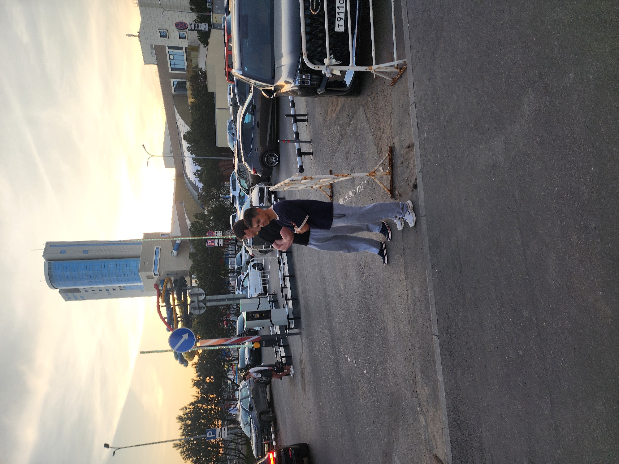 The photograph was taken outdoors during daylight hours. The time of day appears to be dusk or early evening, as indicated by the lighting. Two male individuals are standing on a paved road, possibly a street or parking area. They are both wearing casual clothing, with one in a dark shirt and light-colored pants, and the other in a black shirt and light gray pants.
Several parked cars are visible along the side of the road, some of which are behind a white metal barricade. A modern building can be seen in the background, with architectural features including glass panels and a curved façade. Street signs and traffic control devices are also present.
Based on a street sign and license plate, the location is Kazan, Russia. The license plate on a black SUV shows the regional code for Tatarstan. The photo has been rotated.