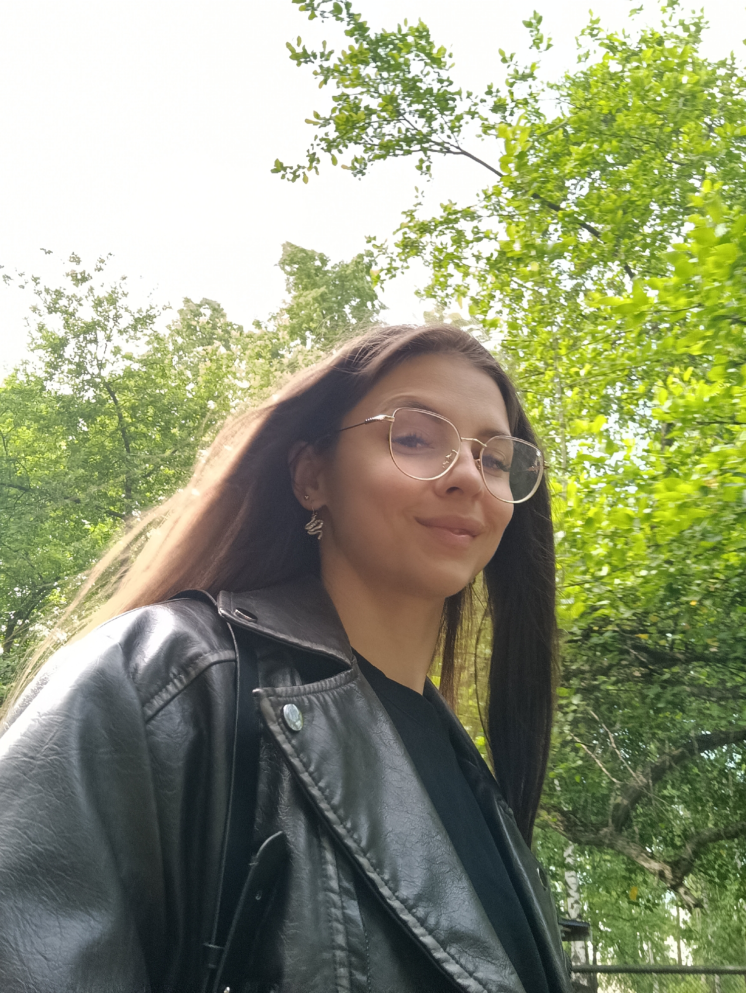 The image is a medium close-up of a young woman outdoors, captured from a slightly low angle. The subject, a woman with long, straight dark brown hair, is positioned in the right half of the frame, looking slightly to her right with a gentle, closed-mouth smile. She wears thin-framed, light-colored glasses and has a serene expression. Her hair appears to be in motion, flowing towards her left, possibly due to a breeze.
She is dressed in a black leather or faux leather jacket over a black top. A small, silver-colored earring with a distinct wavy or snake-like design is visible on her right earlobe.
The background is filled with the vibrant green leaves of numerous trees, suggesting a park, forest, or garden setting. The sky, visible in patches through the tree canopy and dominating the upper left portion of the image, is bright and appears overcast or diffused, indicating daytime. The lighting is soft and natural. No specific location cues beyond "outdoor natural environment" are identifiable, and there is no visible text.