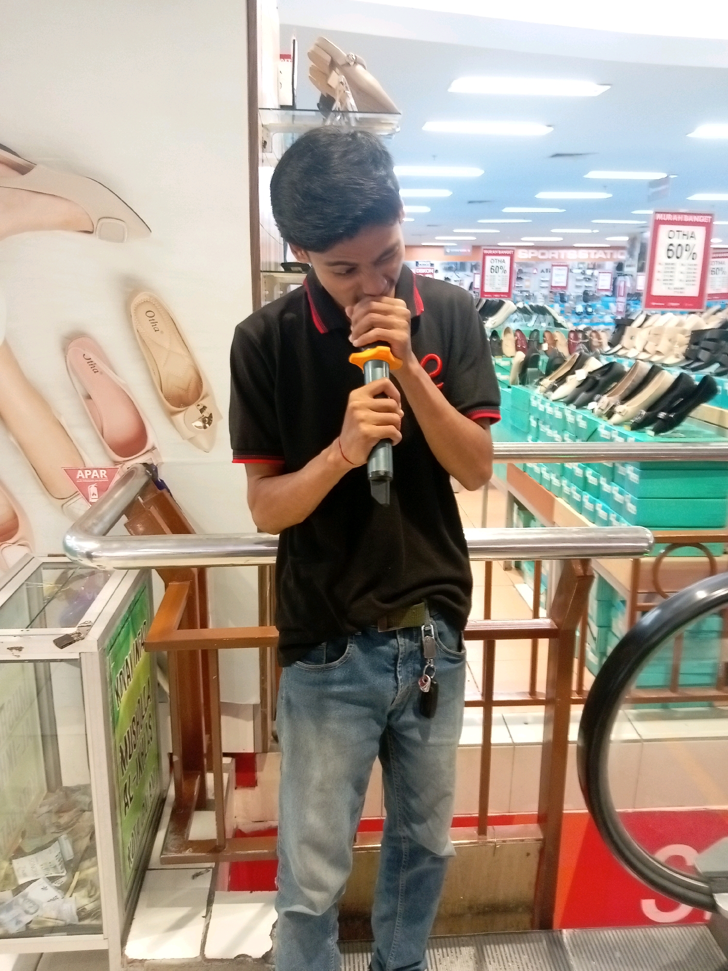 A young male, wearing a black polo shirt with red trim and blue jeans, stands indoors and holds a microphone to his mouth. He appears to be in a shopping mall, possibly in Batam, Indonesia, given the signs with Indonesian text. The background shows displays of shoes and clothing, with overhead lighting and ceiling panels typical of a commercial space. In the foreground, a glass display case with what appears to be cash is visible to the left, and a railing surrounds an escalator further to the right. The overall scene suggests a casual, possibly impromptu, moment within a retail environment.