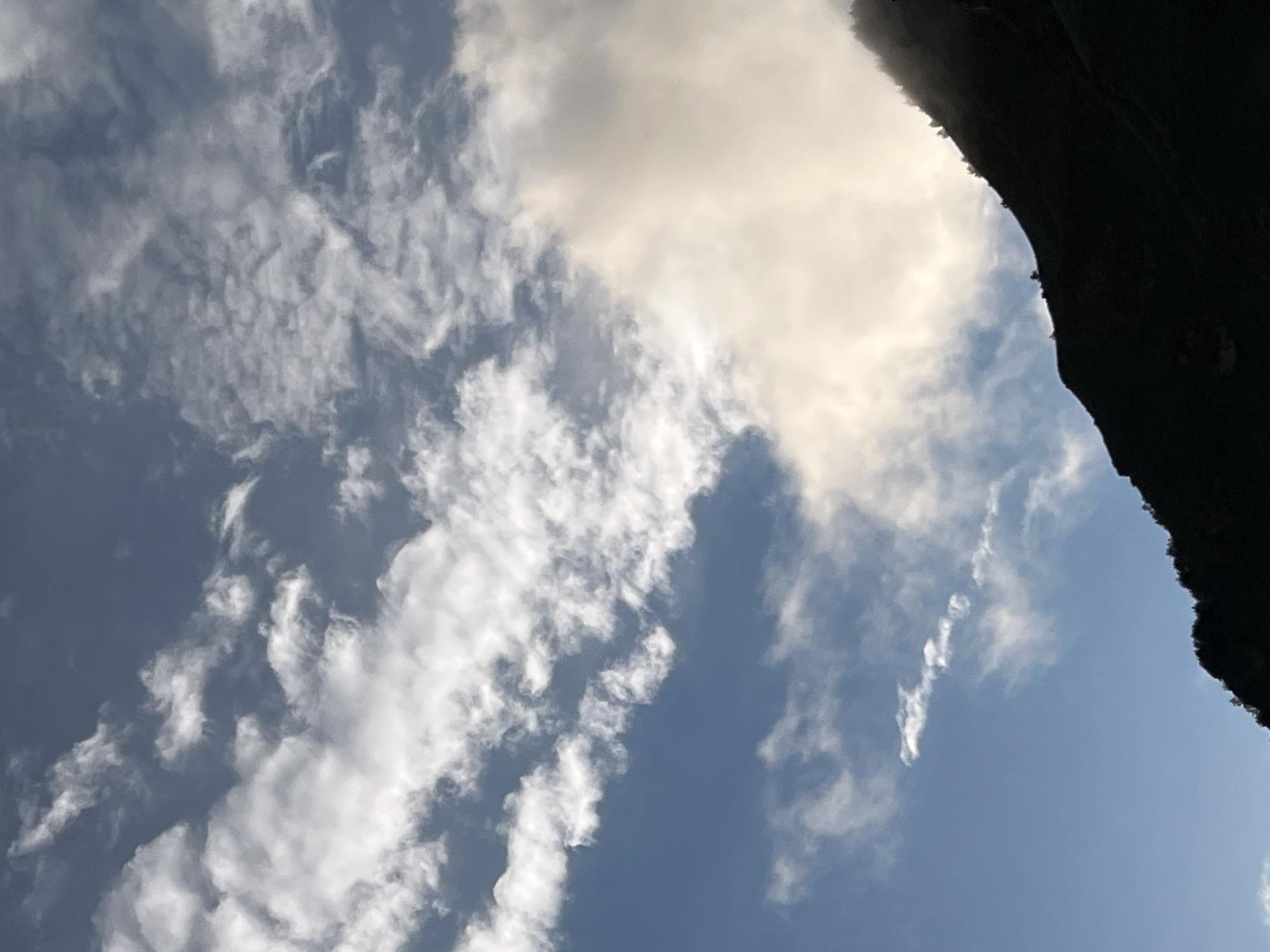 The image shows a silhouetted mountain range against a cloudy sky. The sky is a mix of blue and white clouds, with some areas of bright sunlight peeking through. The mountain range is dark and appears to be covered in trees. The scene suggests an outdoor environment, possibly a natural landscape. There are no people or specific landmarks visible that would indicate a location in the city of Assab, Russia. The image is a photograph capturing a natural vista.