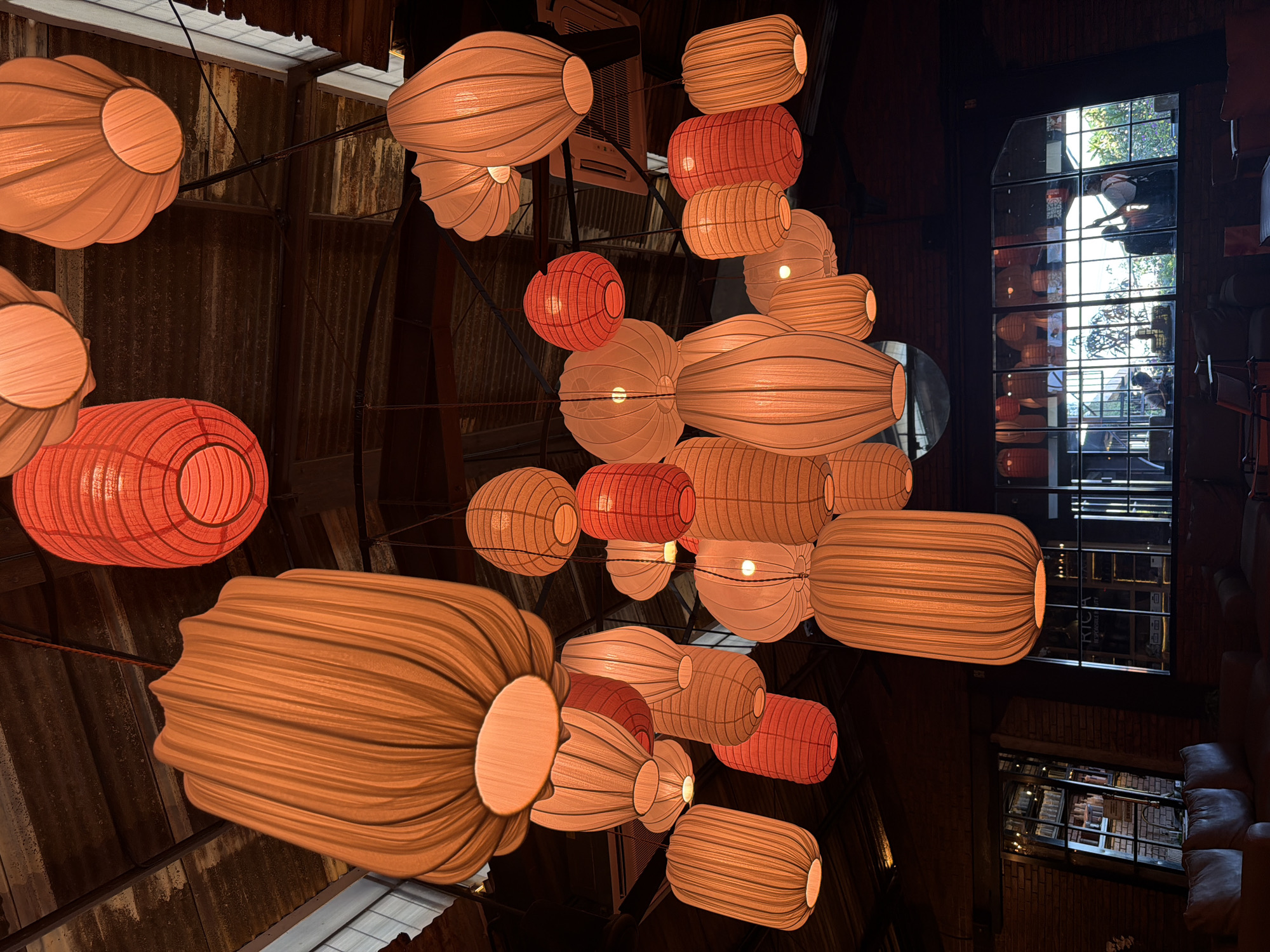 An interior view depicts a ceiling laden with numerous decorative lanterns, primarily orange and beige in color. The lanterns are of varying shapes and sizes, illuminated from within. The ceiling itself appears to be corrugated metal. A large window to the right offers a glimpse outside, reflecting people and possibly the surrounding urban area. Seating, likely a sofa, is visible in the lower right corner of the frame. The image appears to have been taken in North Kuta, Indonesia.