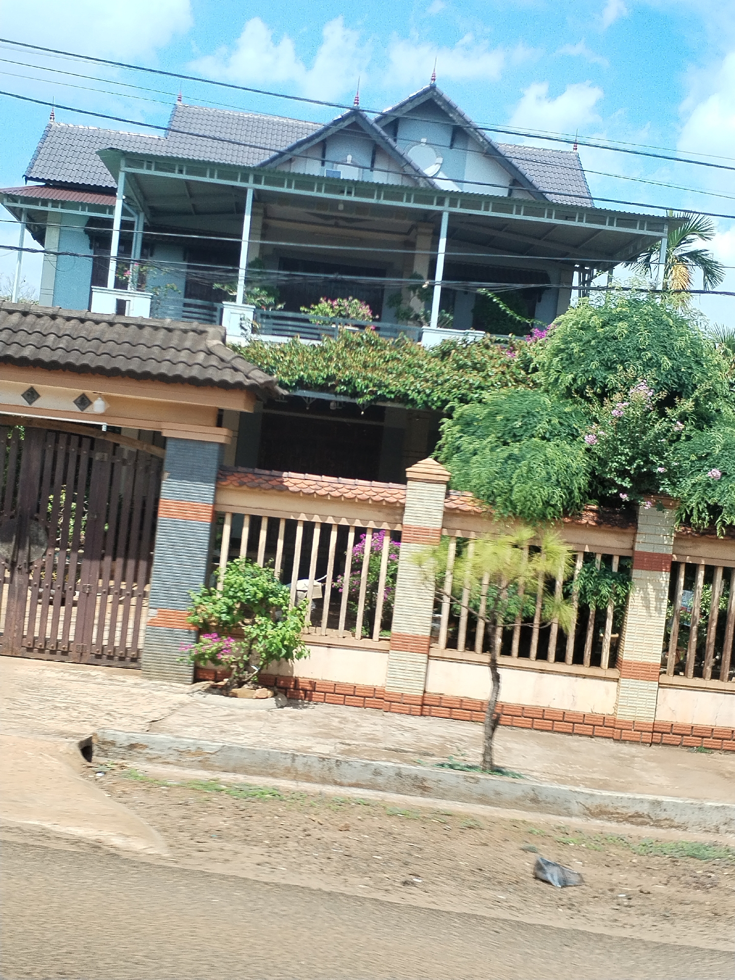 A daytime shot captures a two-story house in Thôn 4, Vietnam. The house features a dark grey tiled roof and a second-floor balcony with a metal frame. Lush green foliage, including flowering plants, drapes over the balcony railing. A wooden fence with brick accents surrounds the property, partially obscured by greenery. A wooden gate stands to the left of the fence. A young tree is planted near the sidewalk. The street in the foreground is paved. Power lines stretch across the sky. The sky is blue with light clouds.