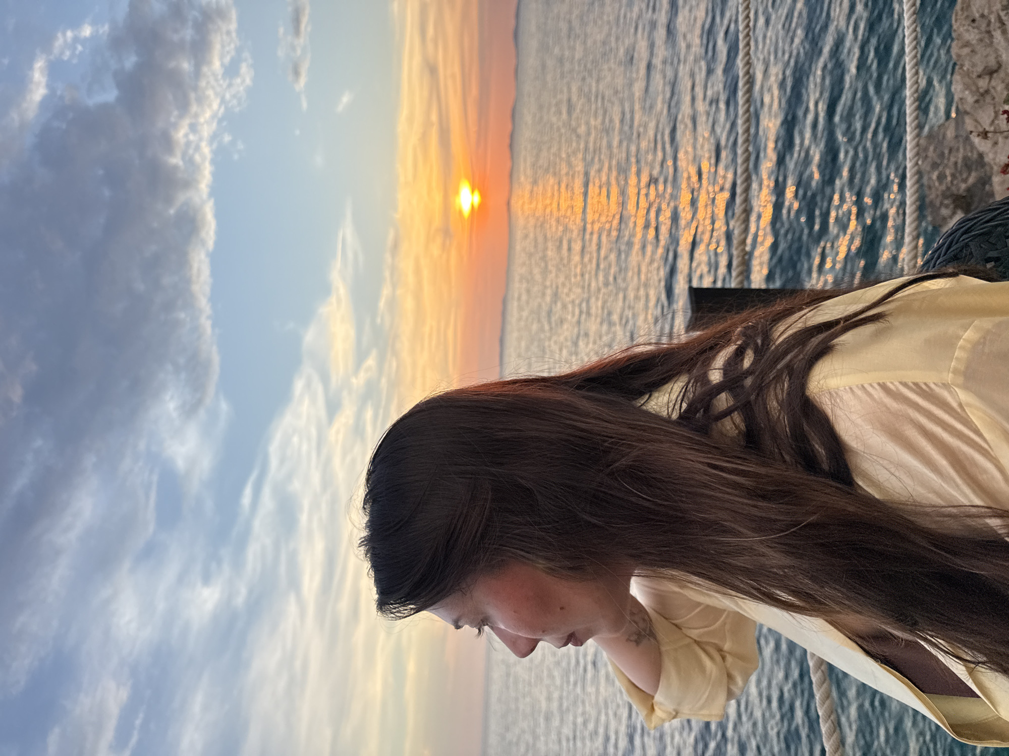 A side-profile medium shot captures a woman with long, dark hair looking out at the sea during sunset in Primošten, Croatia. She is wearing a pale yellow collared shirt, open to reveal a dark garment underneath. Her left hand is resting on the back of her neck, and a tattoo is visible on her arm. The background features the sea, a sunset with orange and yellow hues, and a hazy distant coastline. A rope railing is present in the foreground, and a dark post is visible to the right. The sky is partly cloudy with streaks of white and gray.