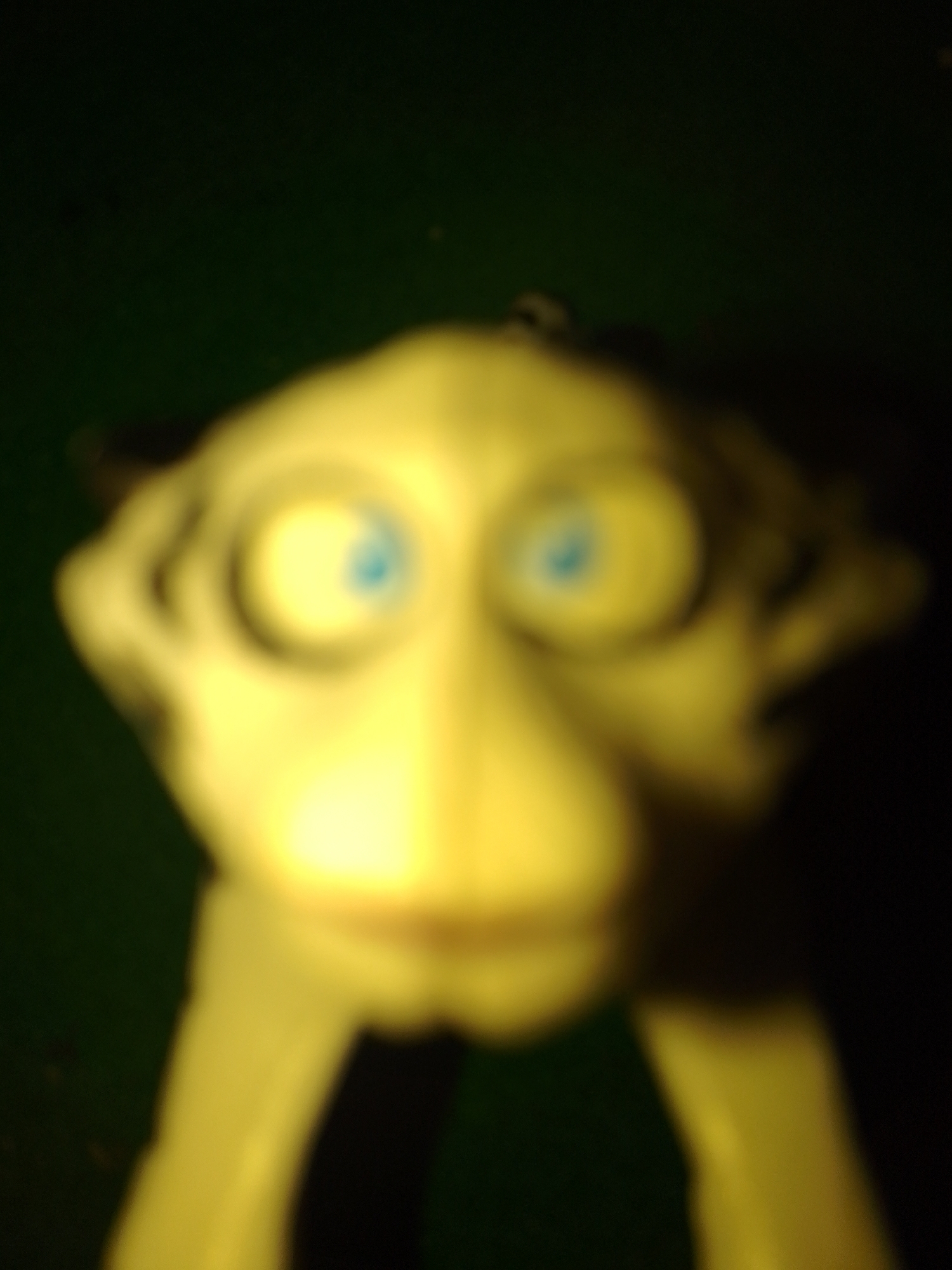 The image is a very blurry, close-up photograph of a toy, likely a figurine. The toy is predominantly yellow and appears to have large, round blue eyes. The background is dark green and out of focus. There is no discernible city of Kauman, Indonesia, or any contextual information that can be derived from the image itself.