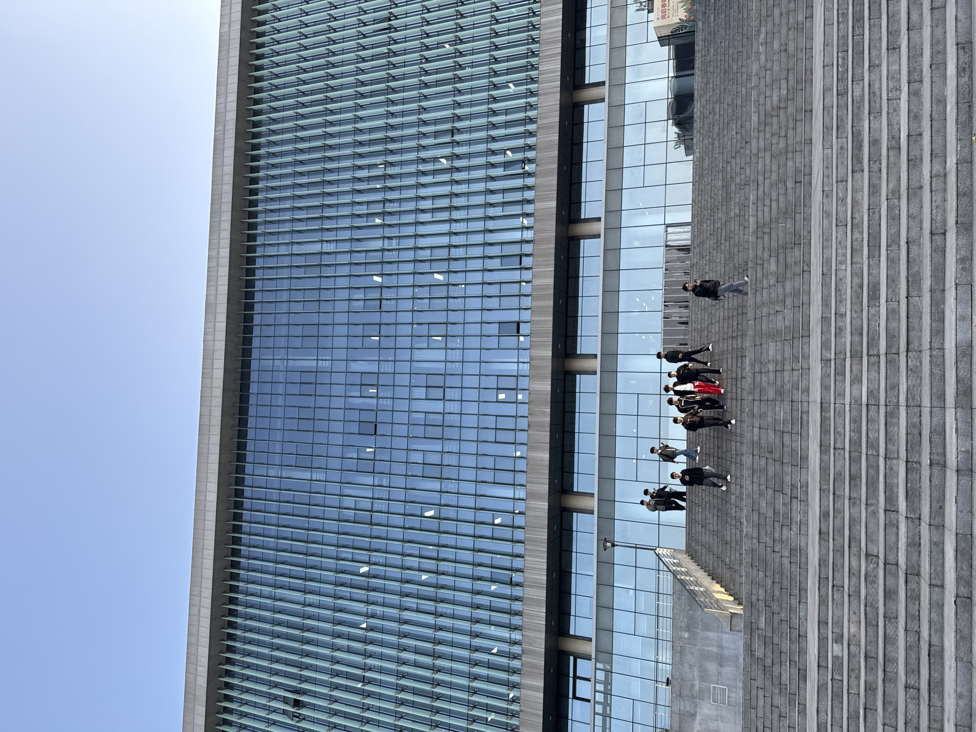 The image depicts a modern building with a glass facade in Jiangjin, China. The left side of the building features a grid pattern of windows with horizontal sunshades, while the right side is a clear glass surface reflecting the surrounding environment. A group of people is standing on a large set of gray concrete steps that run along the base of the building. They appear to be a mix of men and women, some wearing dark jackets and trousers, and one wearing a red jacket. The sky is visible above the building, appearing light blue and clear. The scene appears to be taken at ground level, looking up at the building's exterior.