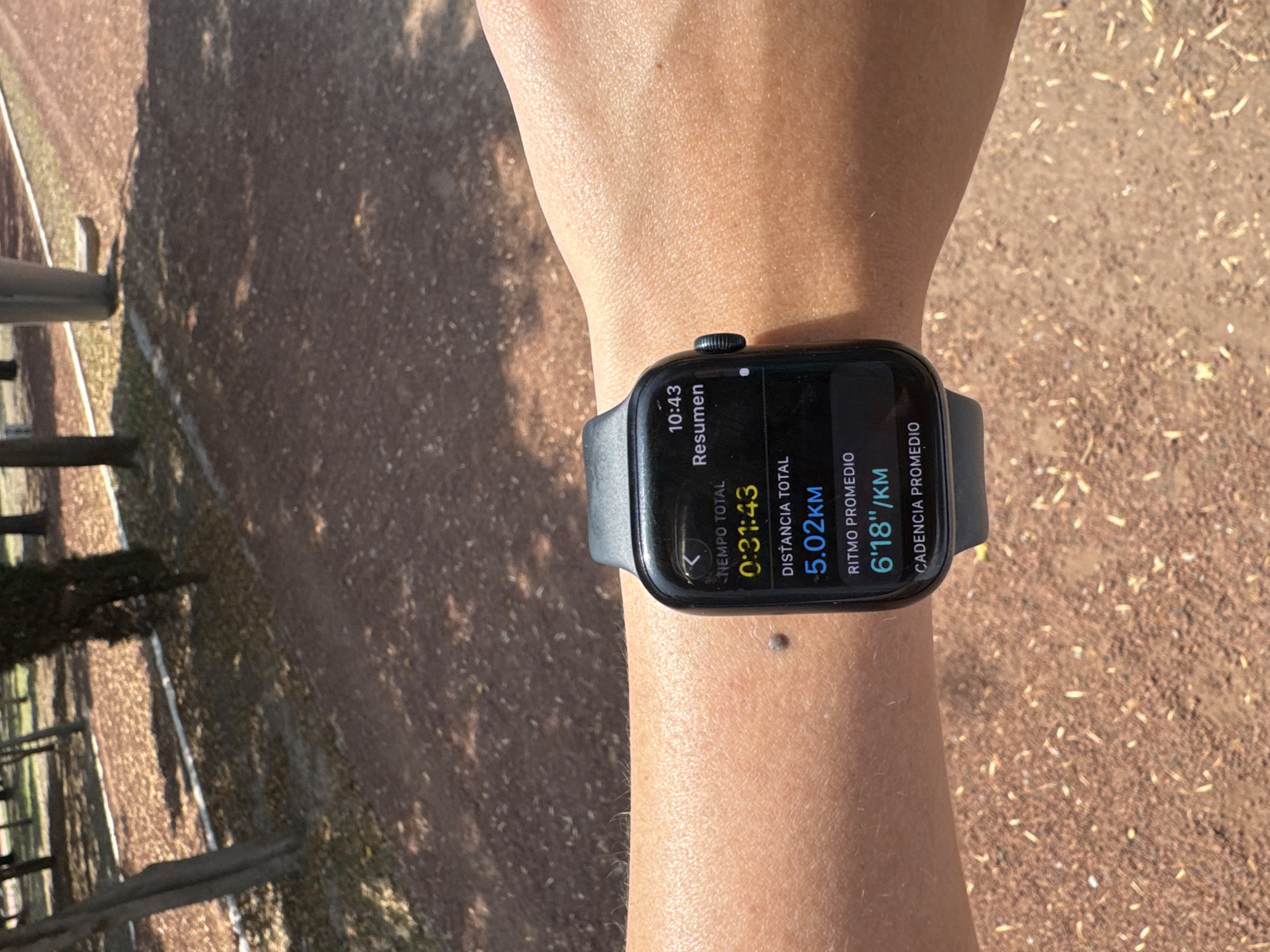 Here is a detailed description of the image:
The image shows a person's wrist wearing a black smartwatch. The watch display reads "10:43 Resumen," indicating it's likely a summary screen. Below that, there is information about an activity: "NEMPO TOTAL 0:31:43" (Total time 0:31:43), "DISTANCIA TOTAL 5.02KM" (Total distance 5.02km), "RITMO PROMEDIO 6'18''/KM" (Average pace 6'18''/km), and "CADENCIA PROMEDIO" (Average cadence). There is a mole on the person's wrist.
The background is a blurred, outdoor environment with reddish-brown ground, tree trunks, and what appear to be rails or posts. The scene suggests a park or track setting, likely in daylight. San Bernardino Tlaxcalancingo, Mexico is not visible.