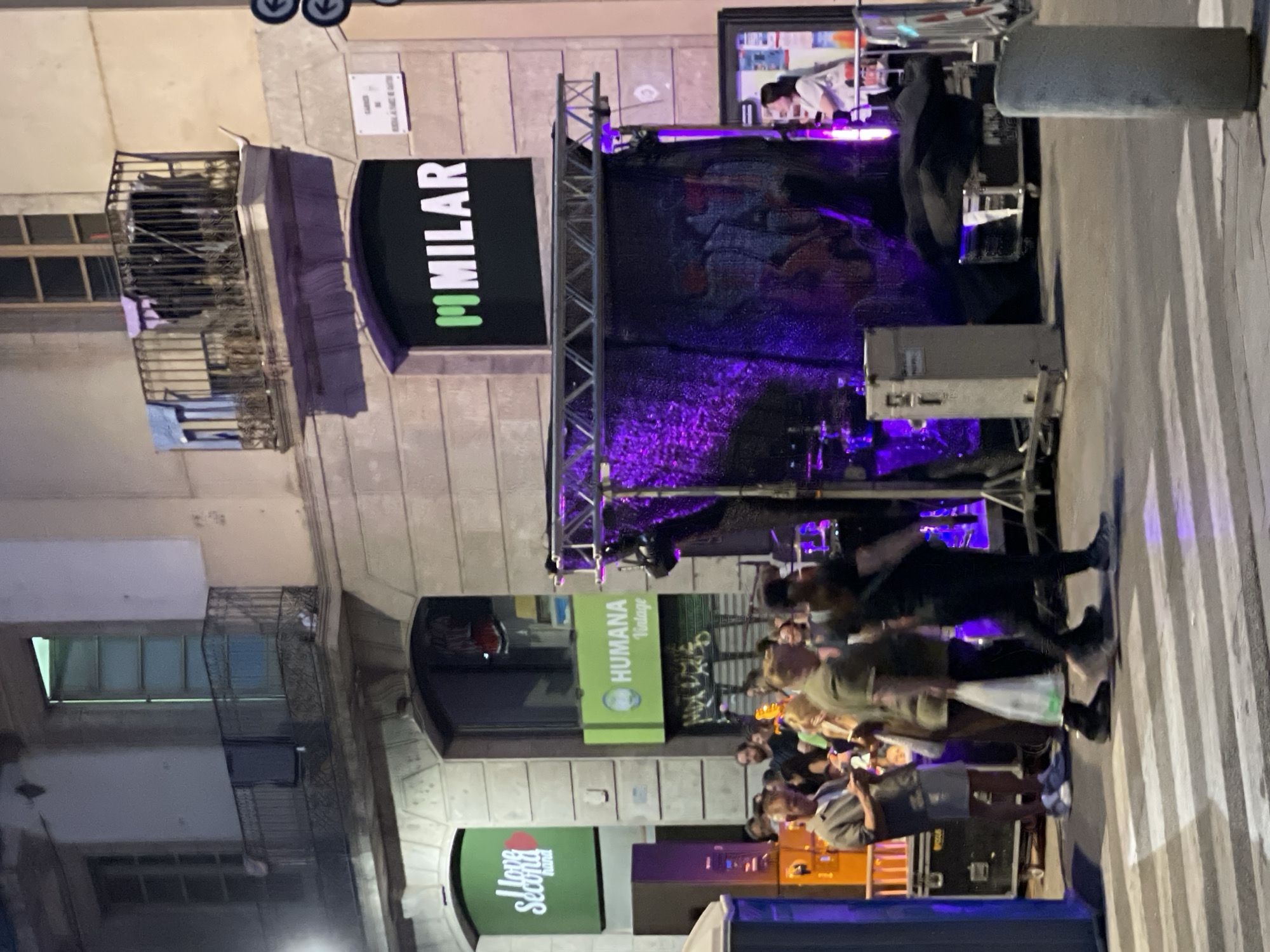 The image depicts a street scene in Barcelona, Spain, likely in the evening, with a group of people gathered near an outdoor stage setup. The backdrop is a building with balconies and windows, some displaying laundry. A prominent sign with "MILAR" in white letters on a black background is visible on the building's facade. Adjacent to it are two green illuminated signs, one reading "I love Second hand" and another "HUMANA Vintage."
In the foreground, several individuals are crossing a pedestrian street, indicated by white stripes. The stage area features lighting equipment, drums, and a dark curtain illuminated with purple light. Several large equipment cases are also present. The overall atmosphere suggests an event or performance taking place outdoors in a commercial or residential area of the city.