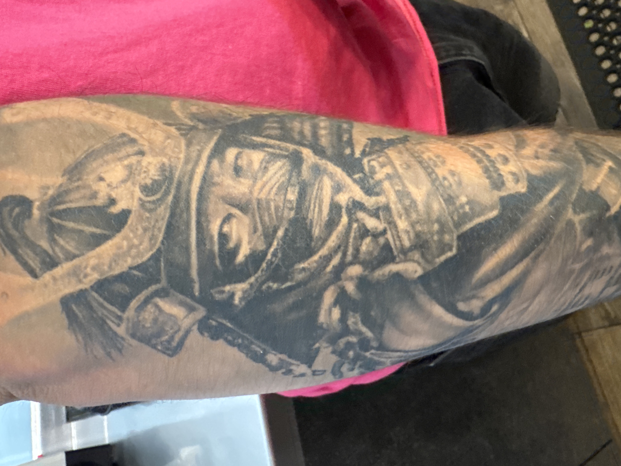 The image shows a close-up of an arm with a detailed black and gray tattoo of a samurai warrior. The arm is light-skinned and slightly hairy. The tattoo covers a significant portion of the forearm, and it appears to be of a high level of detail. The person is wearing a pink shirt or t-shirt on the upper part of the body. In the background, there is a black pair of jeans and a corner of a black keyboard. There is no indication of the city of Provincetown in the image.